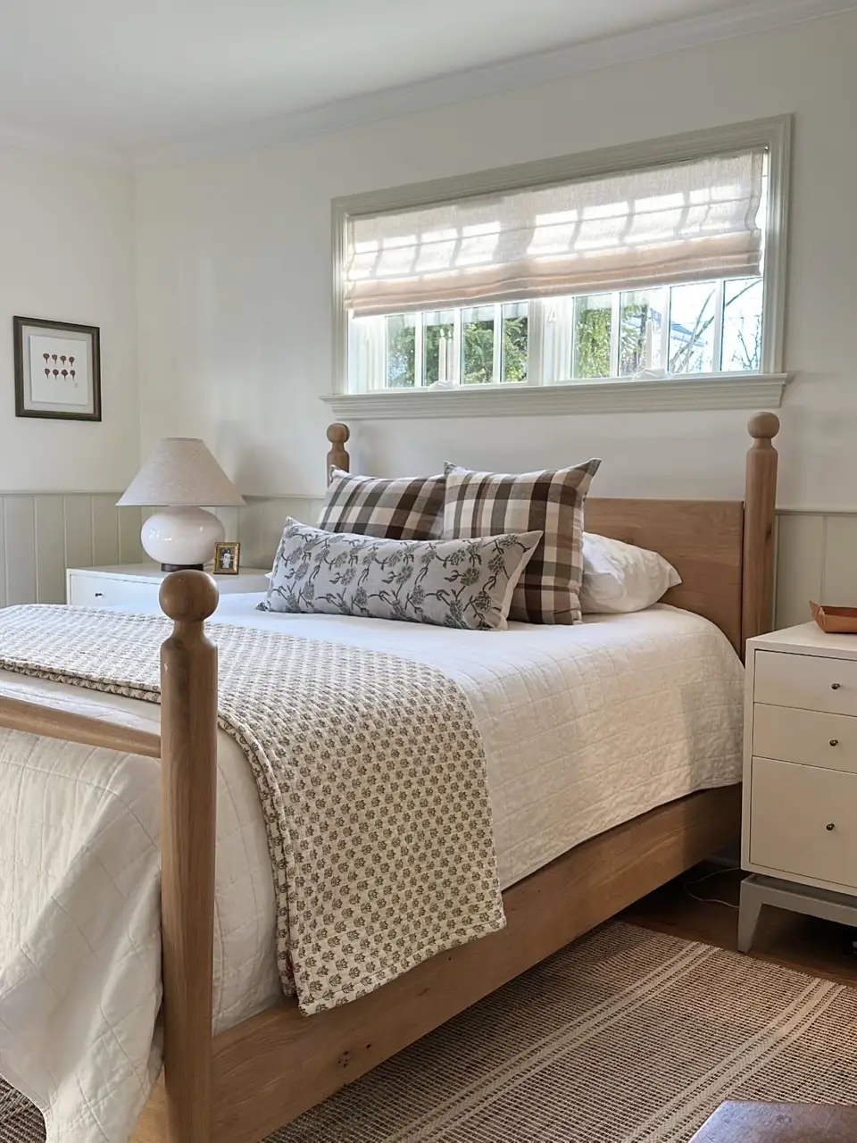 Bright bedroom with a wooden four-poster bed, patterned pillows, white bedding, nightstands, and a window shade.