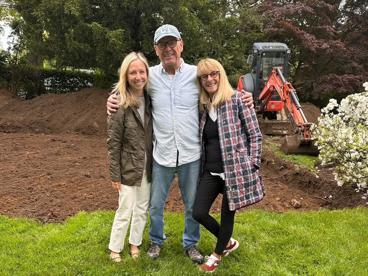 Three people stand on a lawn beside caution tape with an excavator and fresh soil behind them.