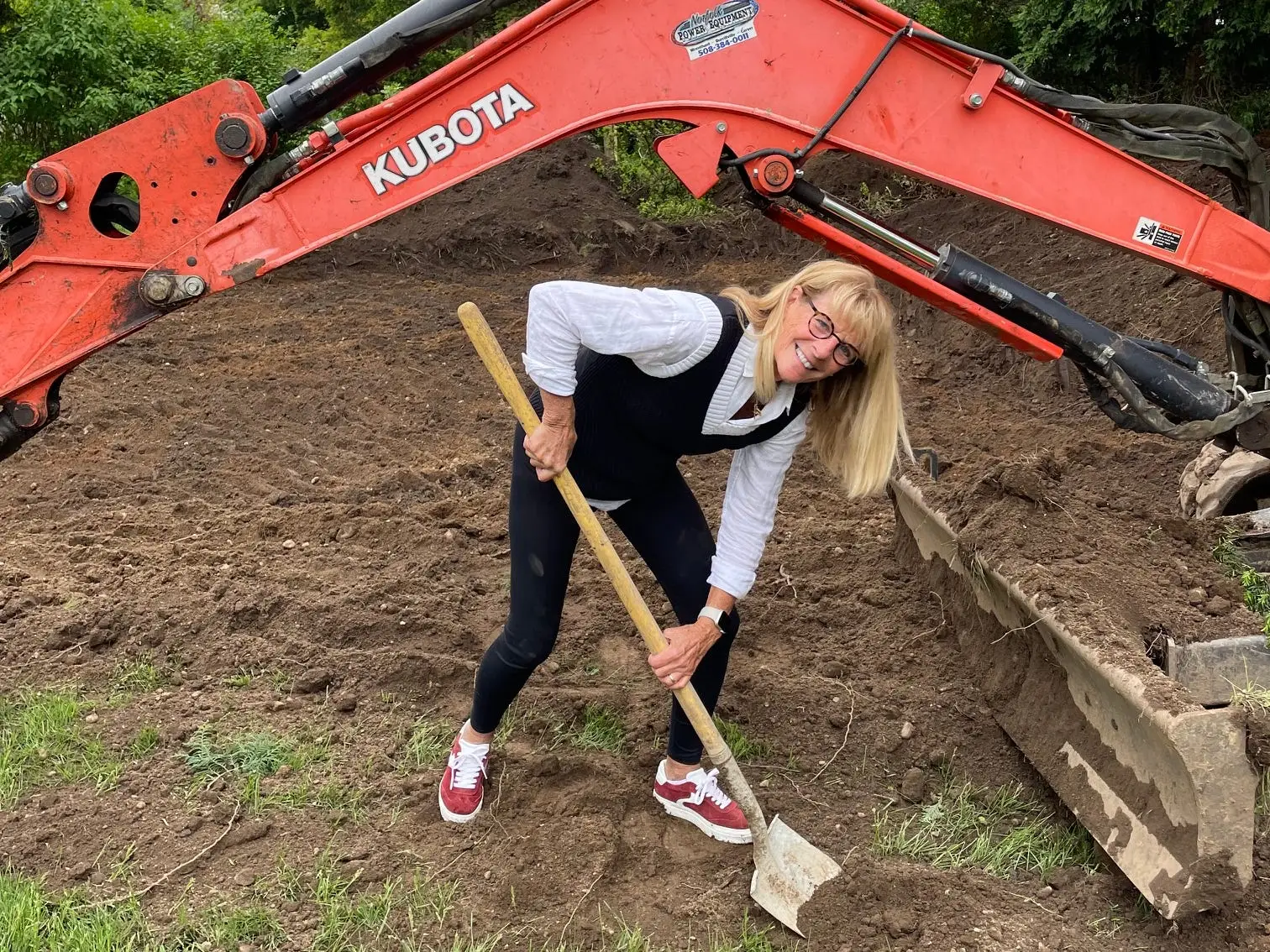 A woman shovels soil beside a red KUBOTA excavator arm in a tree-lined yard.