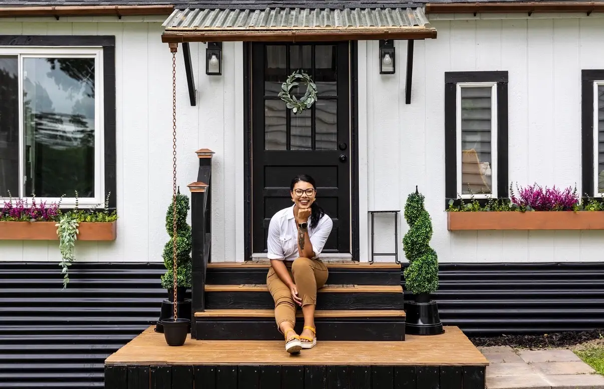 Person sits smiling on front steps of a small white house with black trim, planters, and wreath.