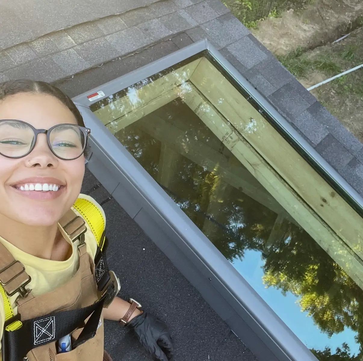 Smiling person in glasses and safety harness stands on a shingled roof beside a newly installed skylight.