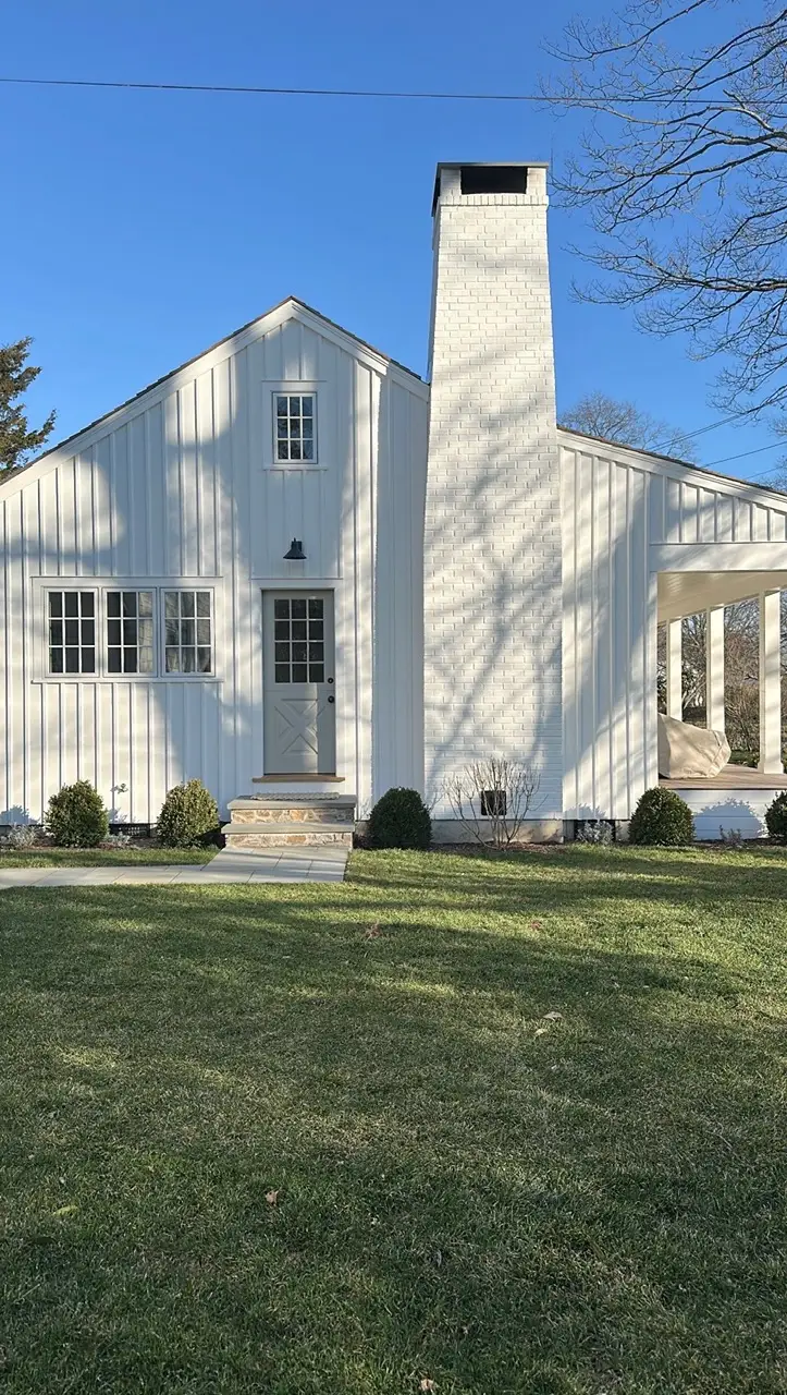 White house exterior with tall brick chimney, front door, porch, shrubs, and lawn under a clear blue sky.