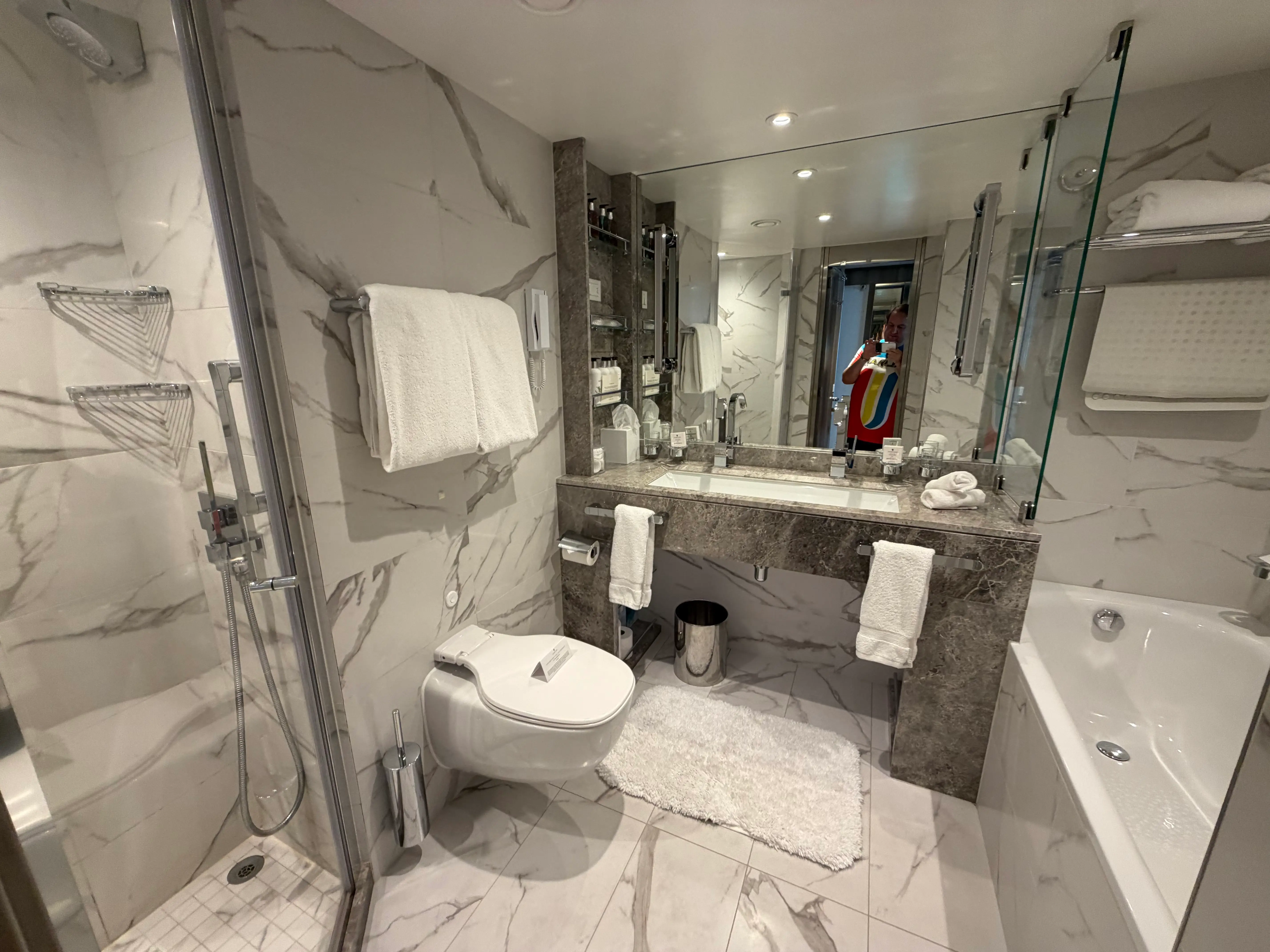 White marble bathroom with shower, large mirror, toilet, vanity ar