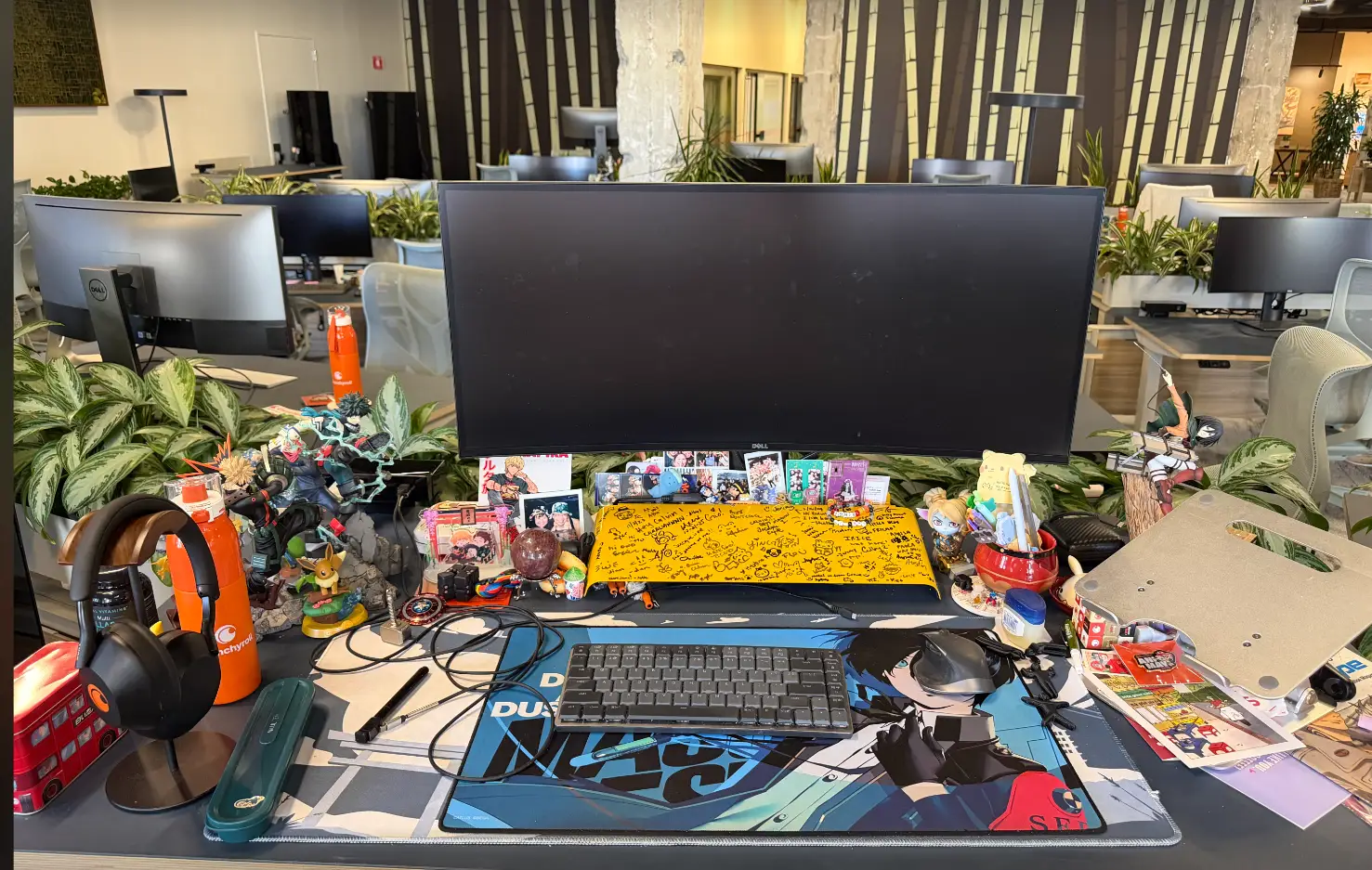 Cluttered office workstation with a Dell monitor, keyboard, anime figures, headphones, plants, and collectibles on a shared desk.