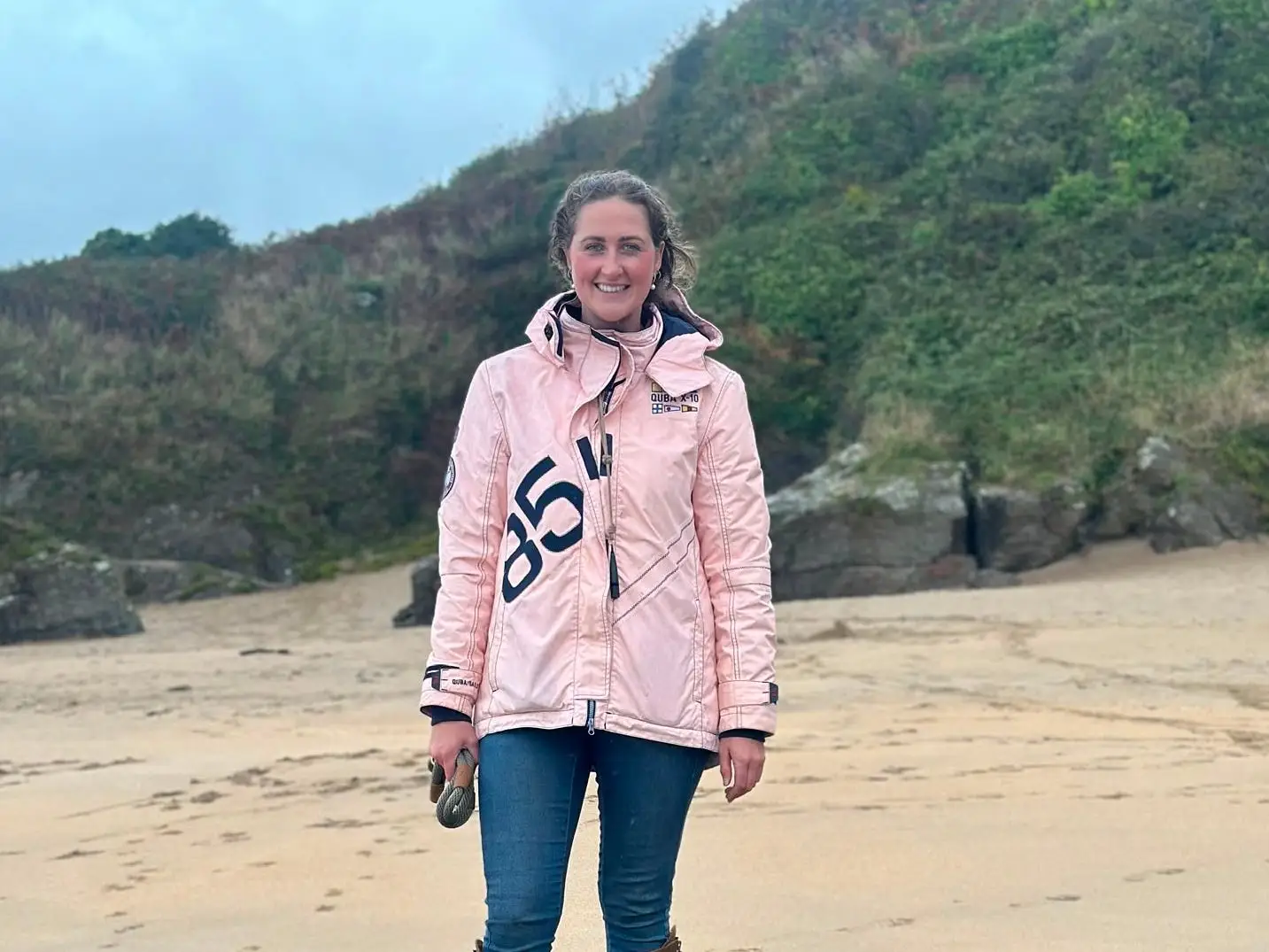 A person in a pink jacket stands with a black dog on rocks at a sandy beach.