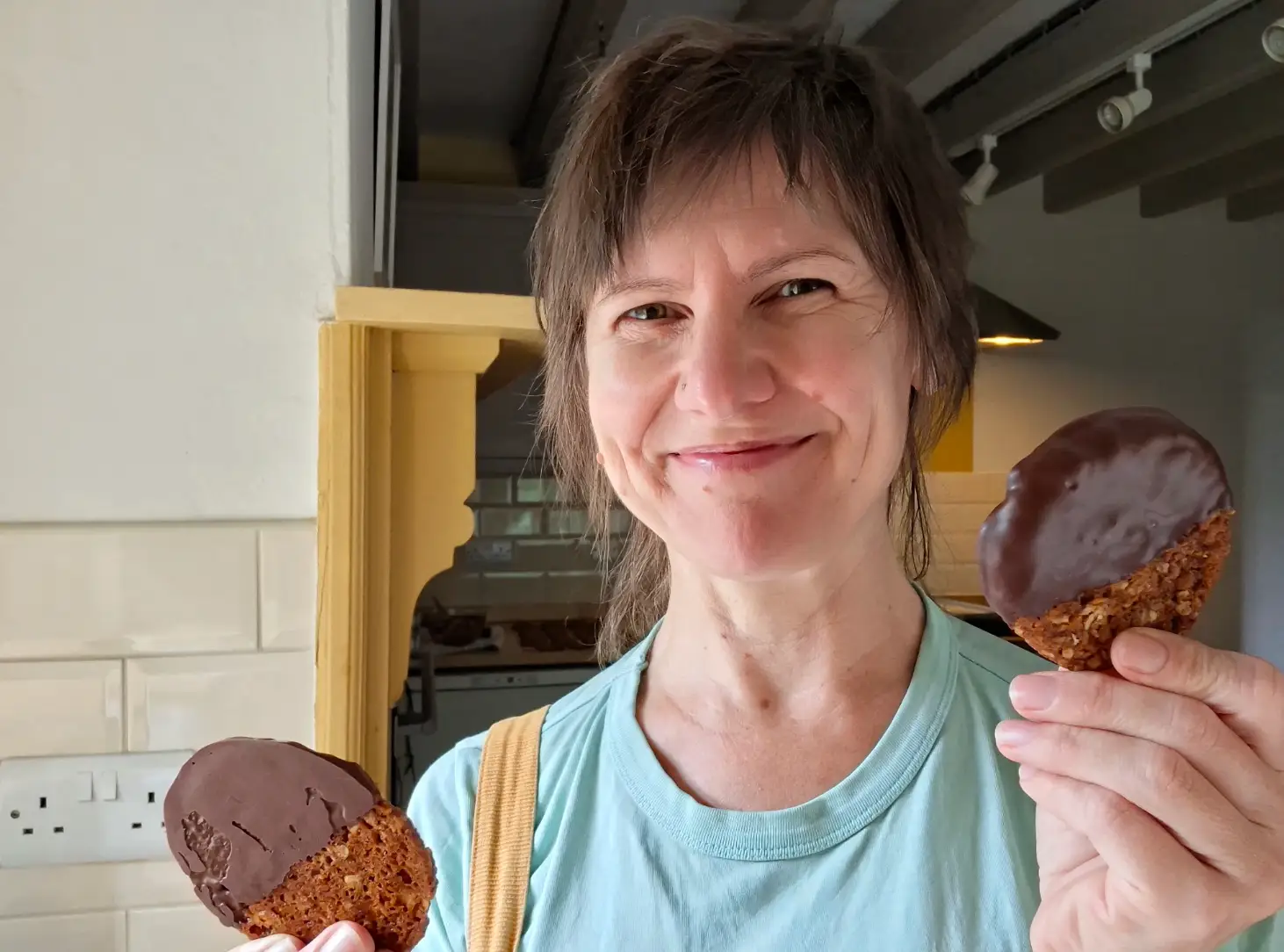 The writer holding two chocolate-dipped cookies and wearing a blue shirt.