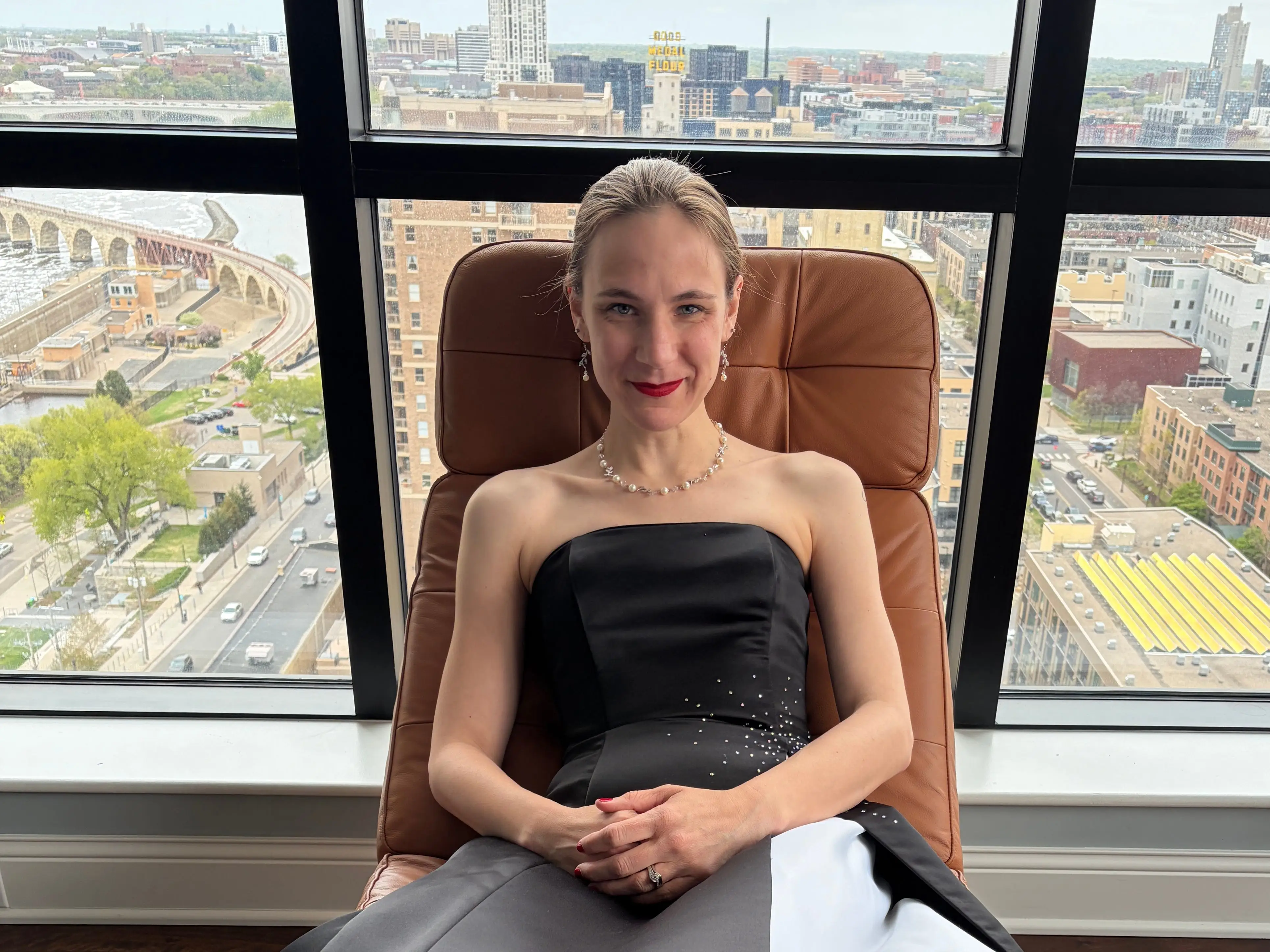 Woman in gown smiling in leather chair in front of large windows with view of city