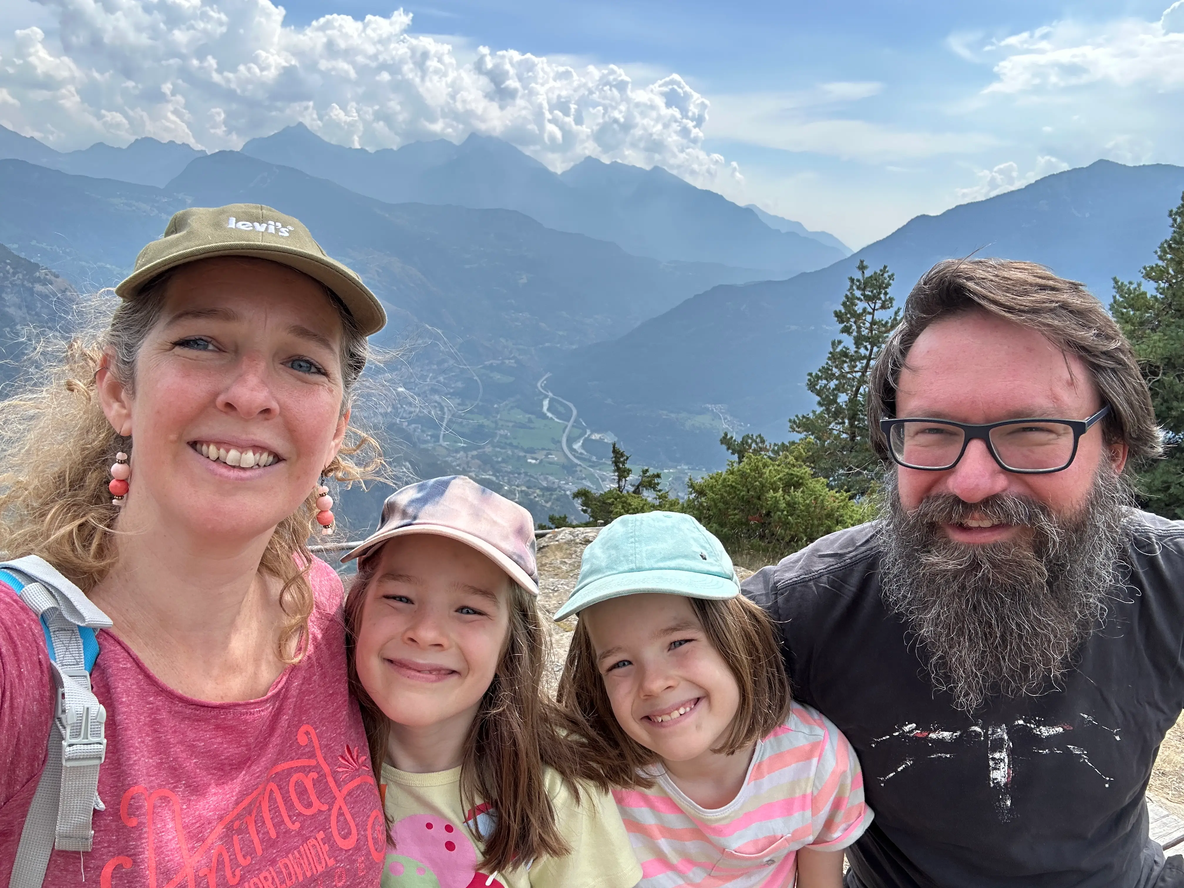Four smiling people pose for a selfie at a mountain overlook with a valley and clouds behind them.
