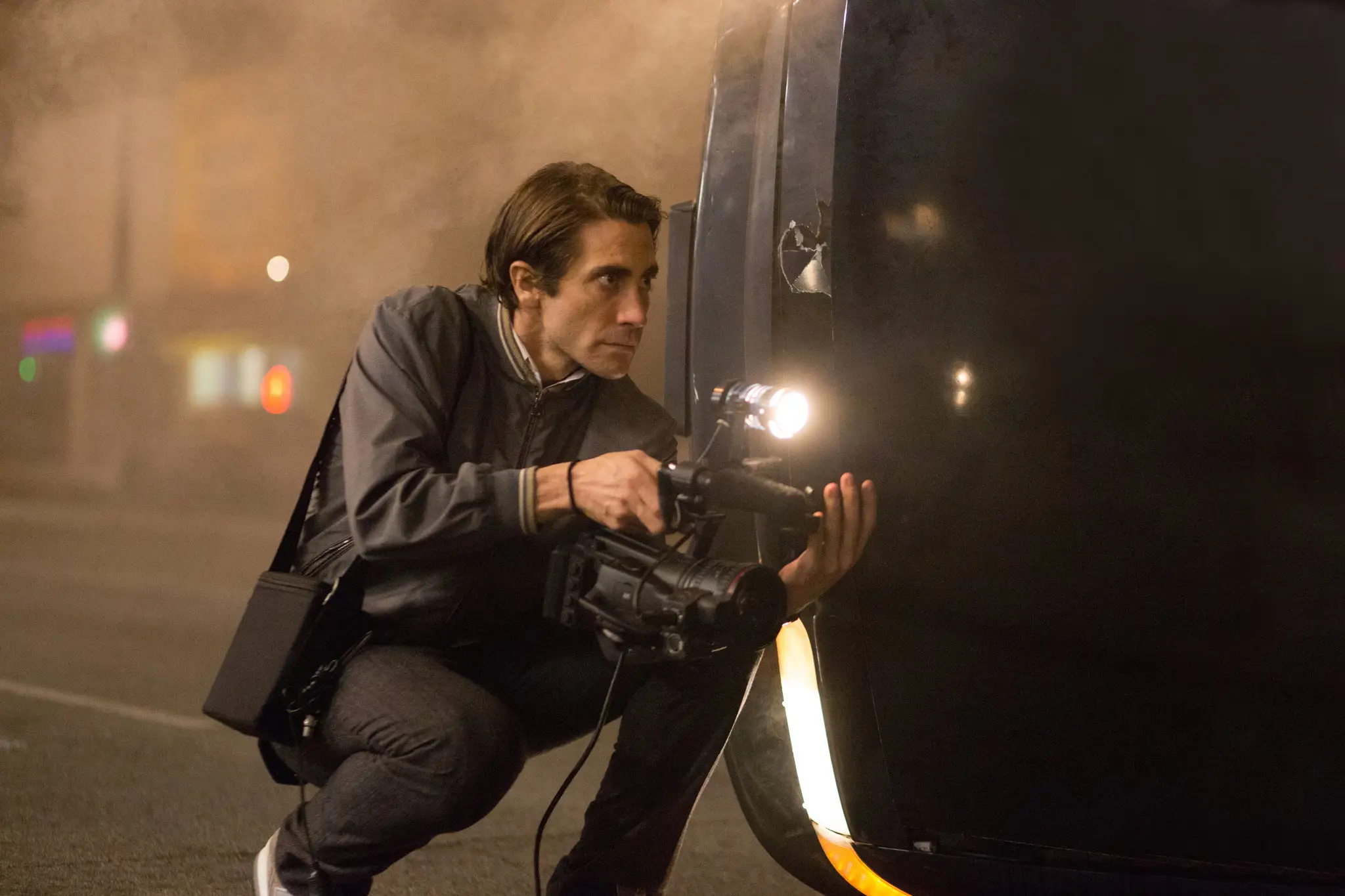 Jake Gyllenhaal holding a camera kneeling next to an overturned car