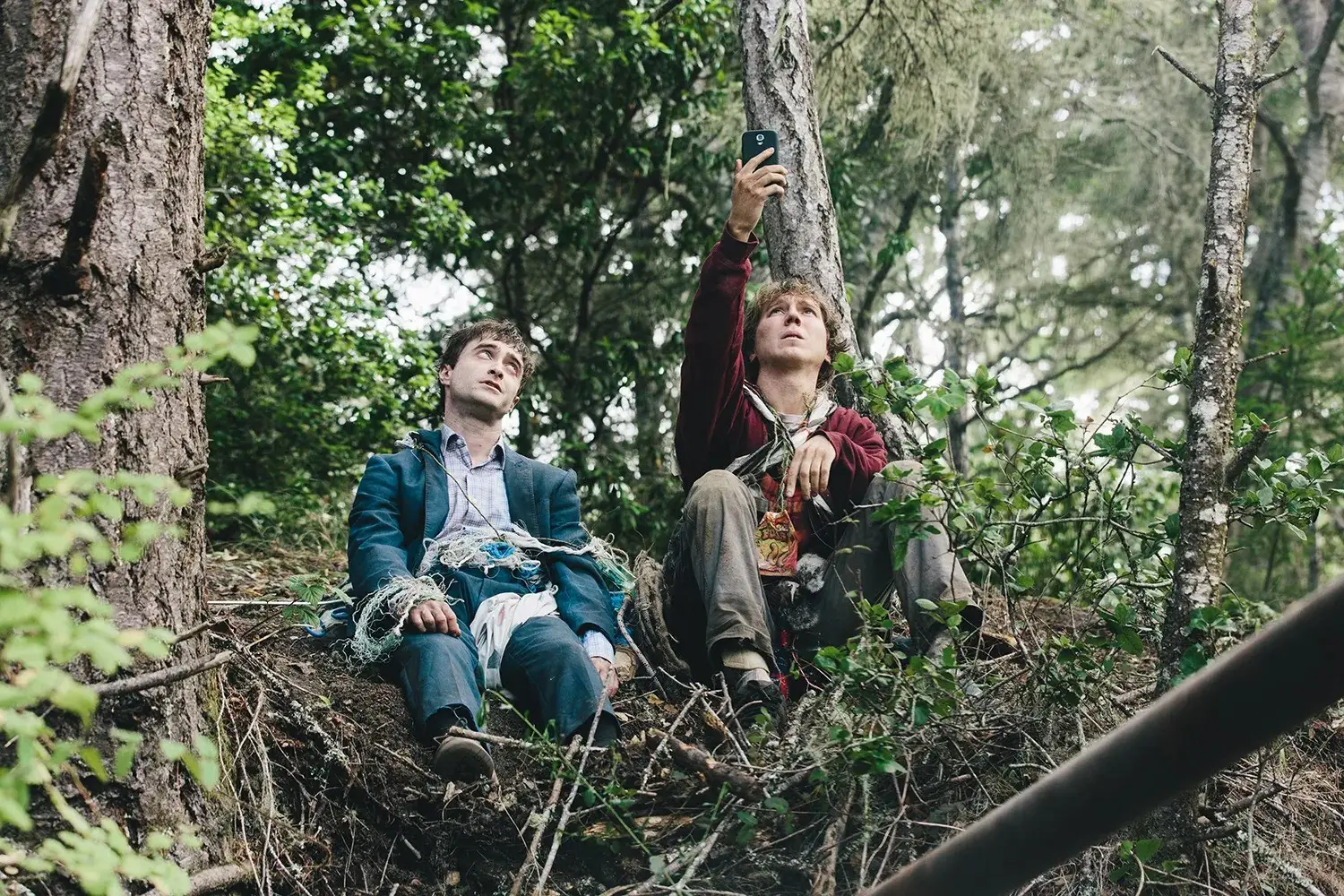 Daniel Radcliffe and Paul Dano sitting in the woods; Dano holing up his phone for reception