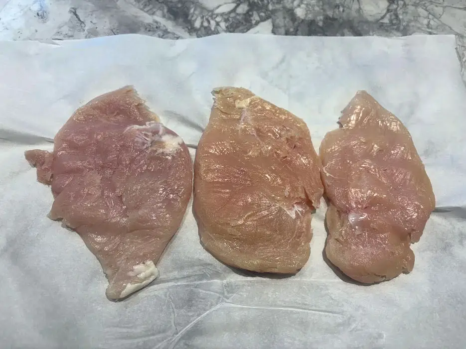 Three flattened raw chicken cutlets on a piece of wax paper.