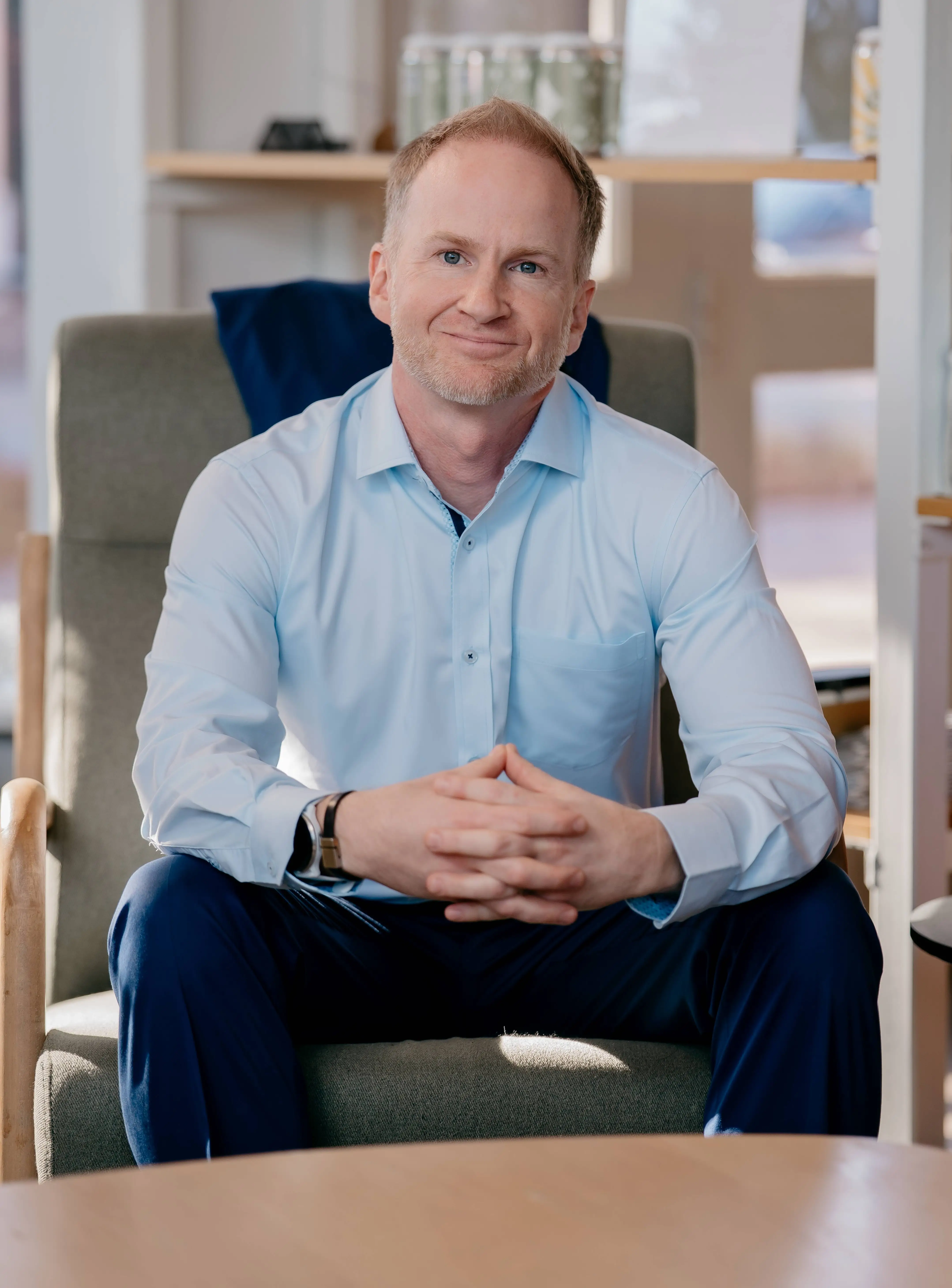 Seated person in a light blue shirt poses with hands clasped in a modern office lounge.