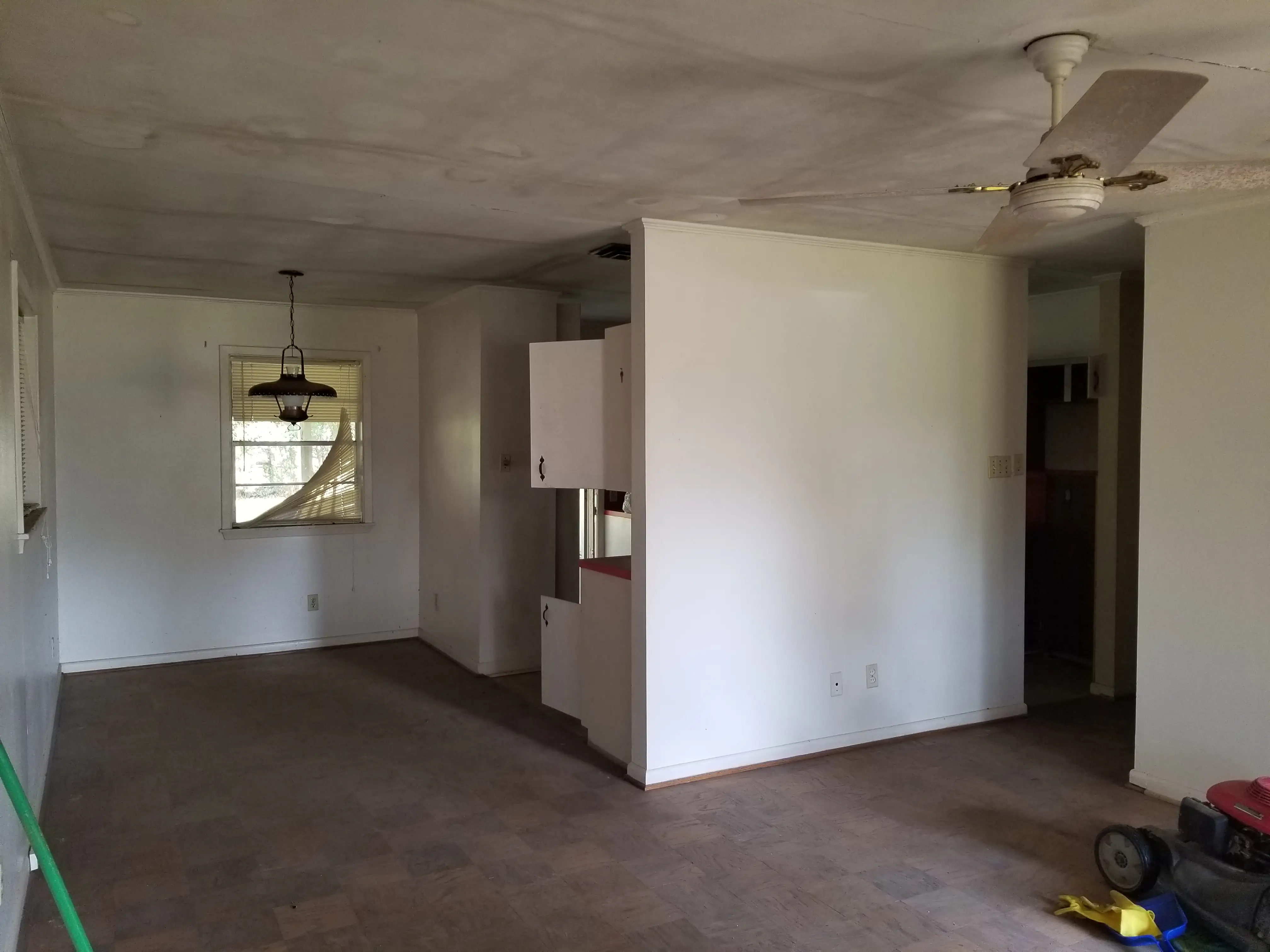 Worn empty interior room with white walls, ceiling fan, damaged window blind, cabinets, and a lawn mower on the floor.