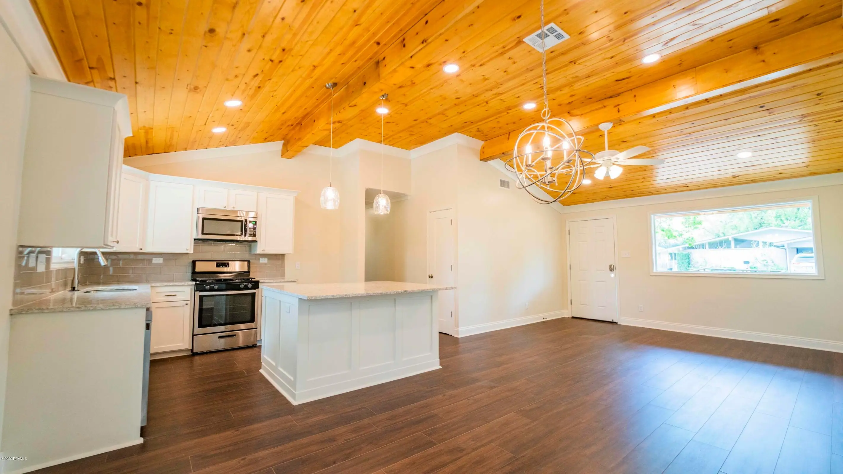 Open-plan kitchen and living area with white cabinets, stainless appliances, wood flooring, and a vaulted wood ceiling.