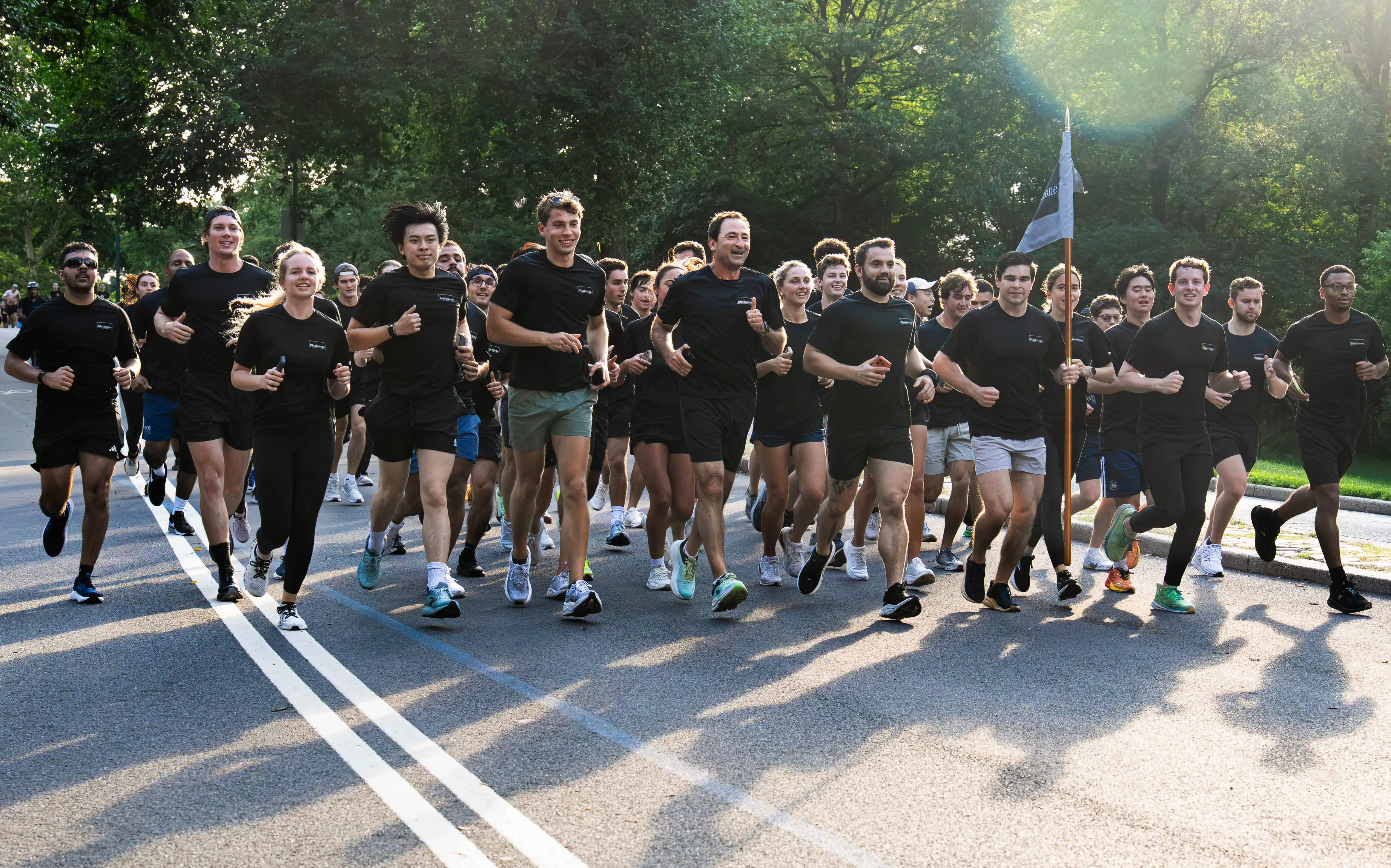 Large group of runners in black shirts jog together on a tree-lined road in bright sunlight.