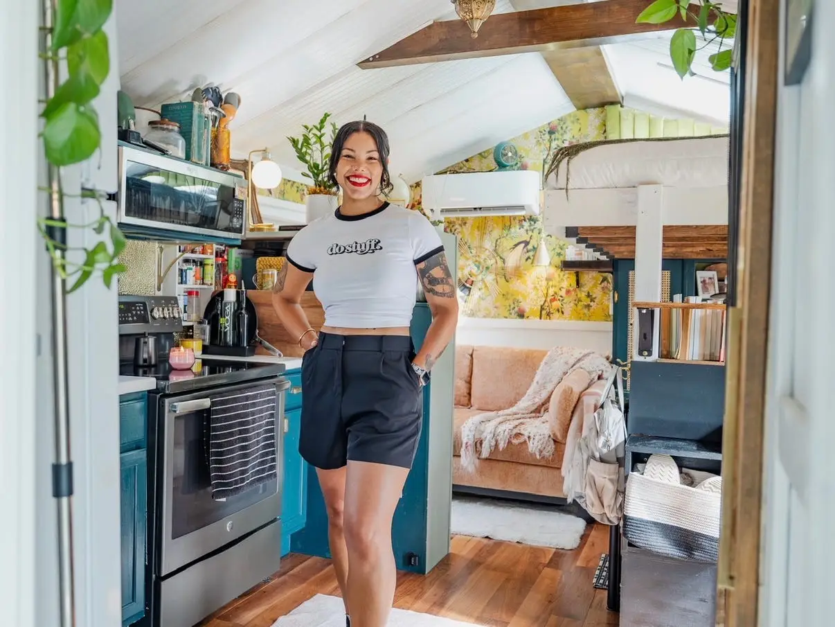 Margo Fairchild stands in a tiny home kitchen with plants, a loft bed, a couch, and wood floors in the background.