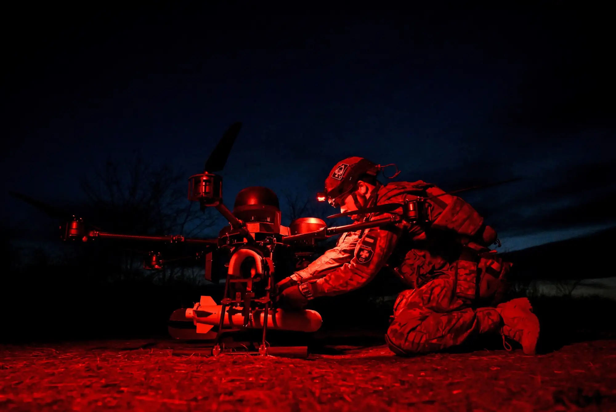 Uniformed person kneels beside a large quadcopter drone under red light during a nighttime outdoor operation.
