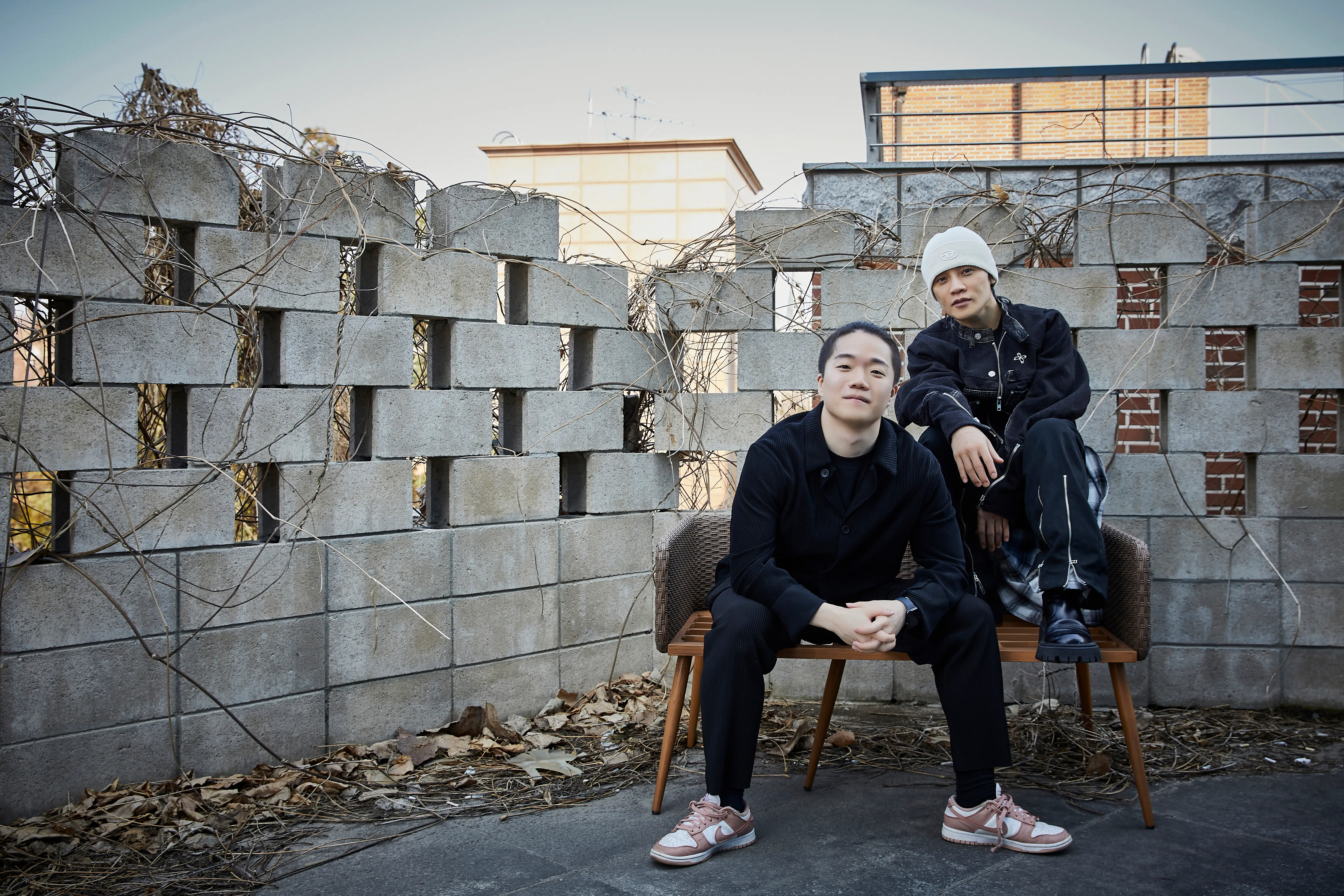 Two people pose on a bench in front of a concrete block wall with bare vines outdoors.