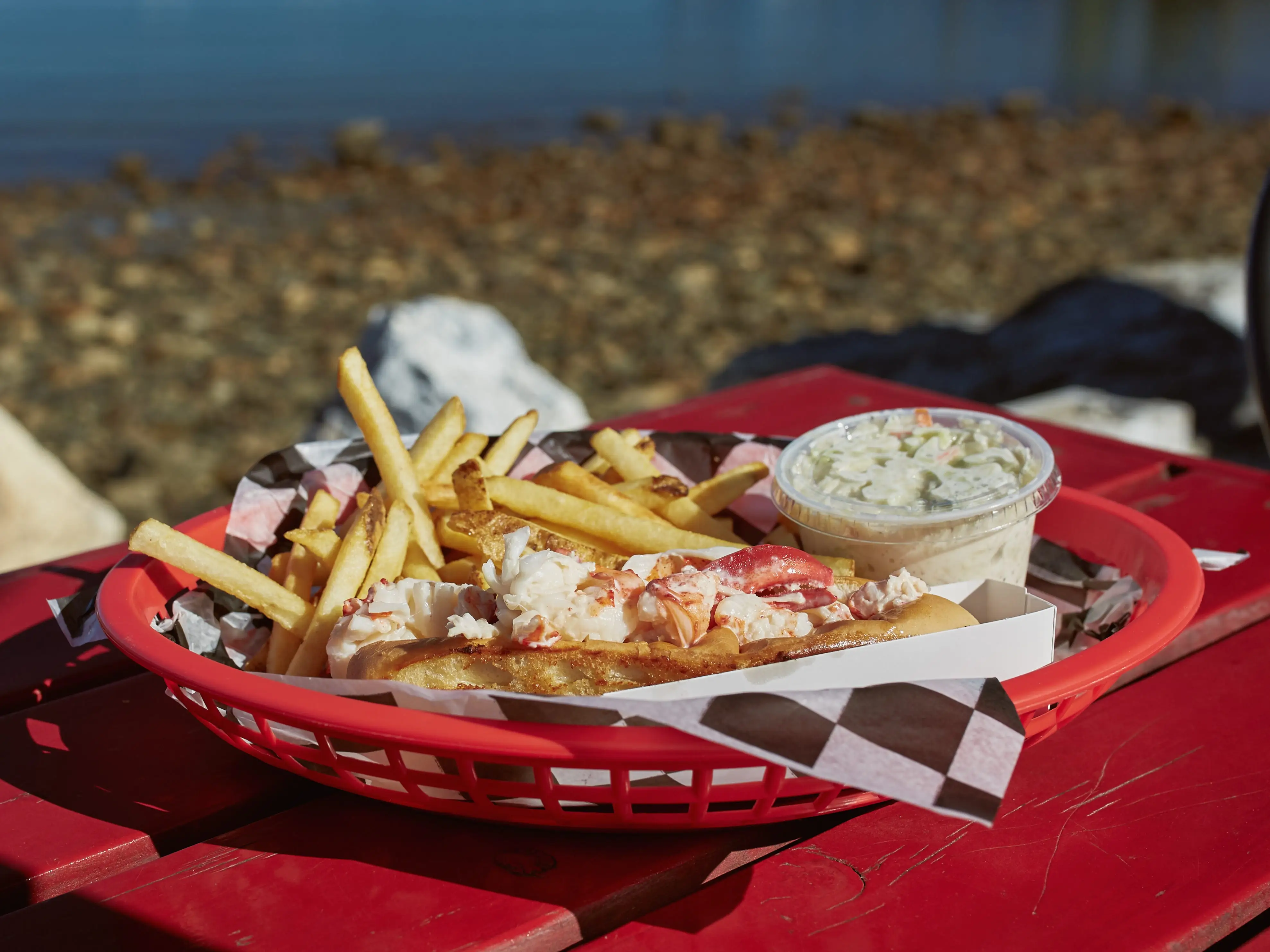 Lobster roll with fries and coleslaw sits in a red basket on a waterfront picnic table.