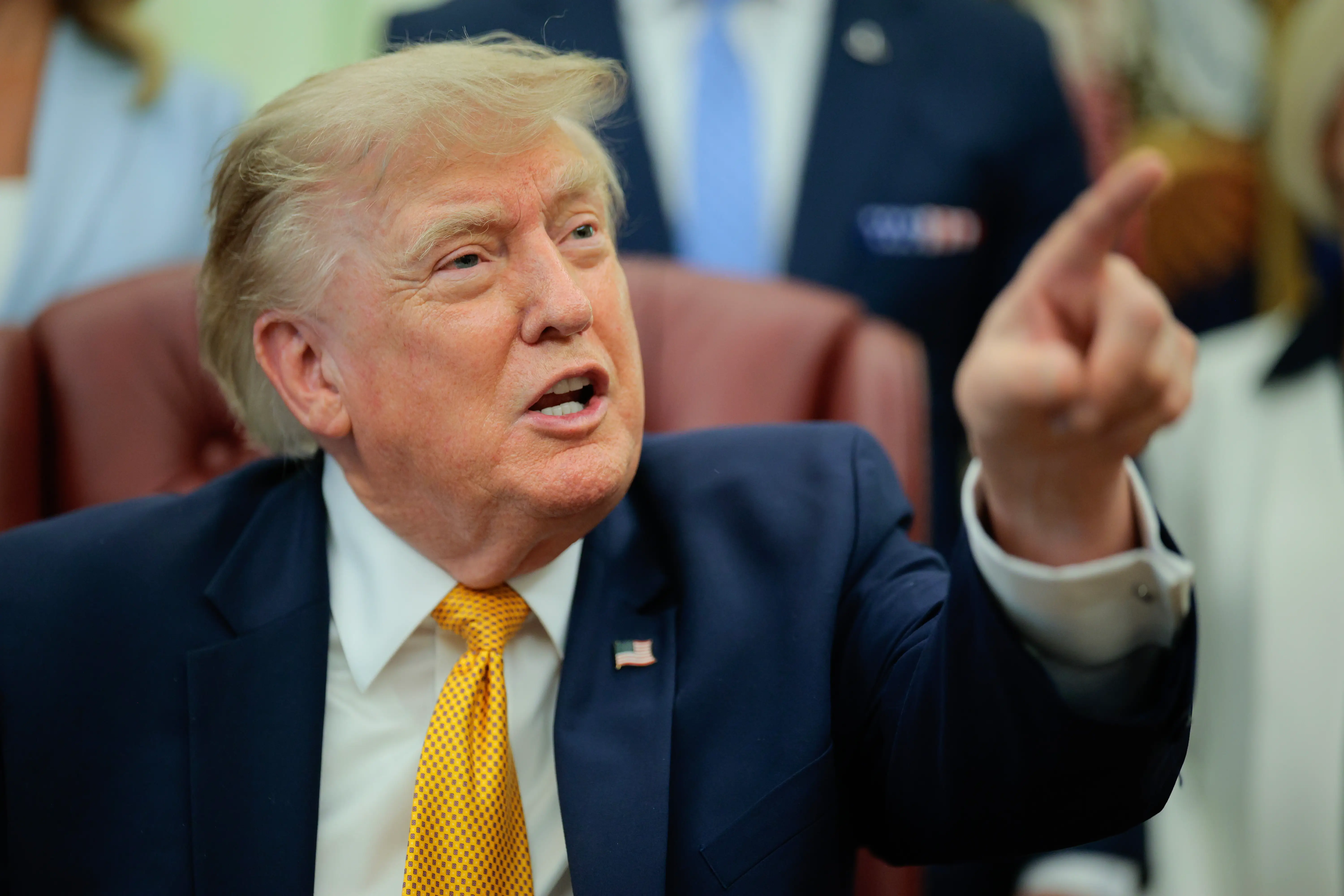 Donald Trump sits in a red chair while speaking and pointing across an office setting.