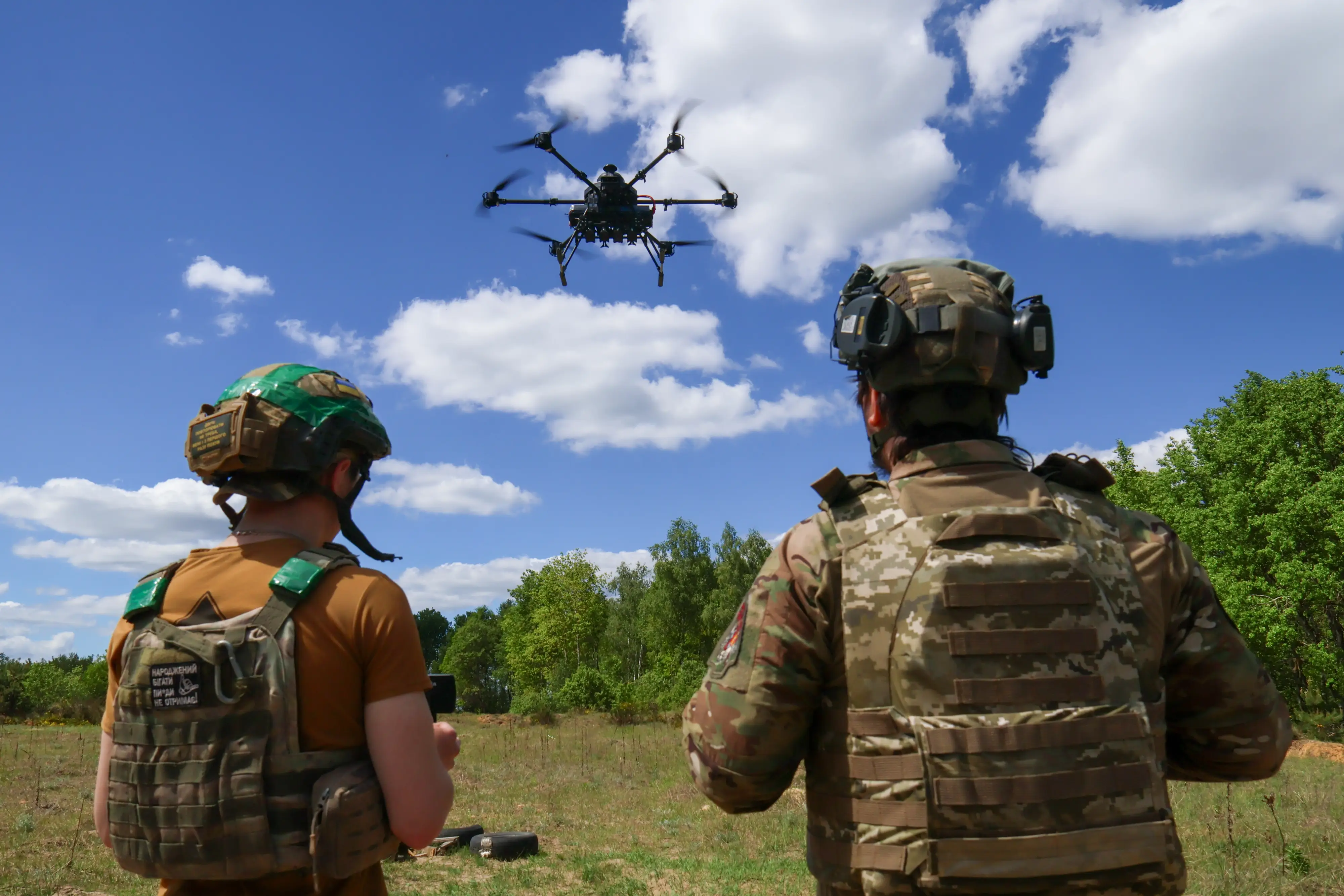 Two uniformed personnel in tactical gear stand in a field while a quadcopter drone flies overhead.