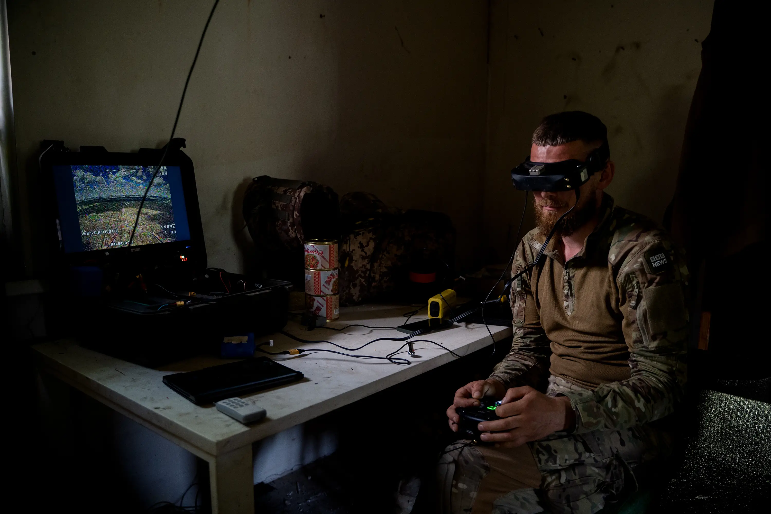 Person in camouflage operates FPV goggles and a controller beside a monitor in a dimly lit room.