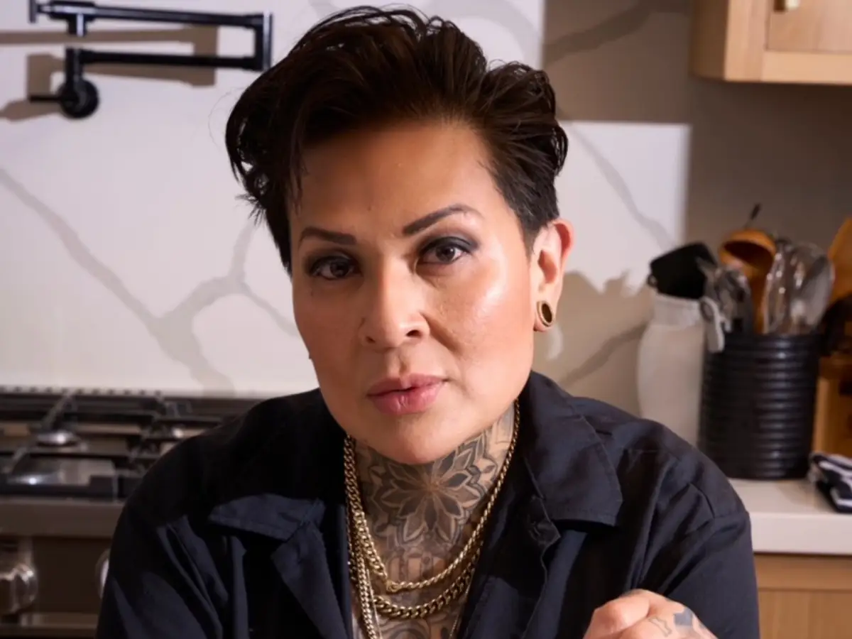 Chef K faces the camera in a kitchen with a stove, utensils, dark shirt, gold chains, and visible tattoos.