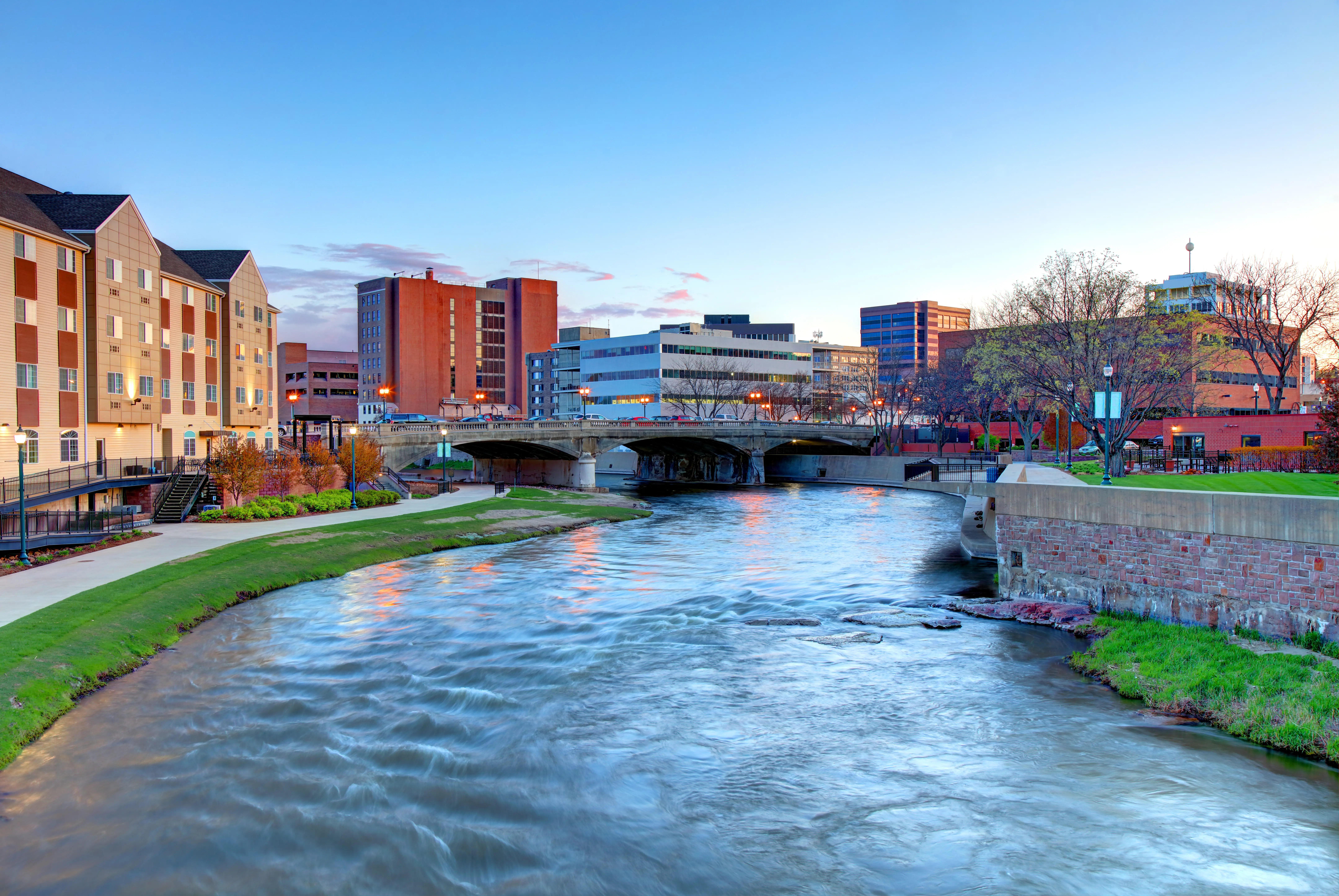 Urban river flows beneath a bridge between downtown buildings at dusk under a clear blue sky.