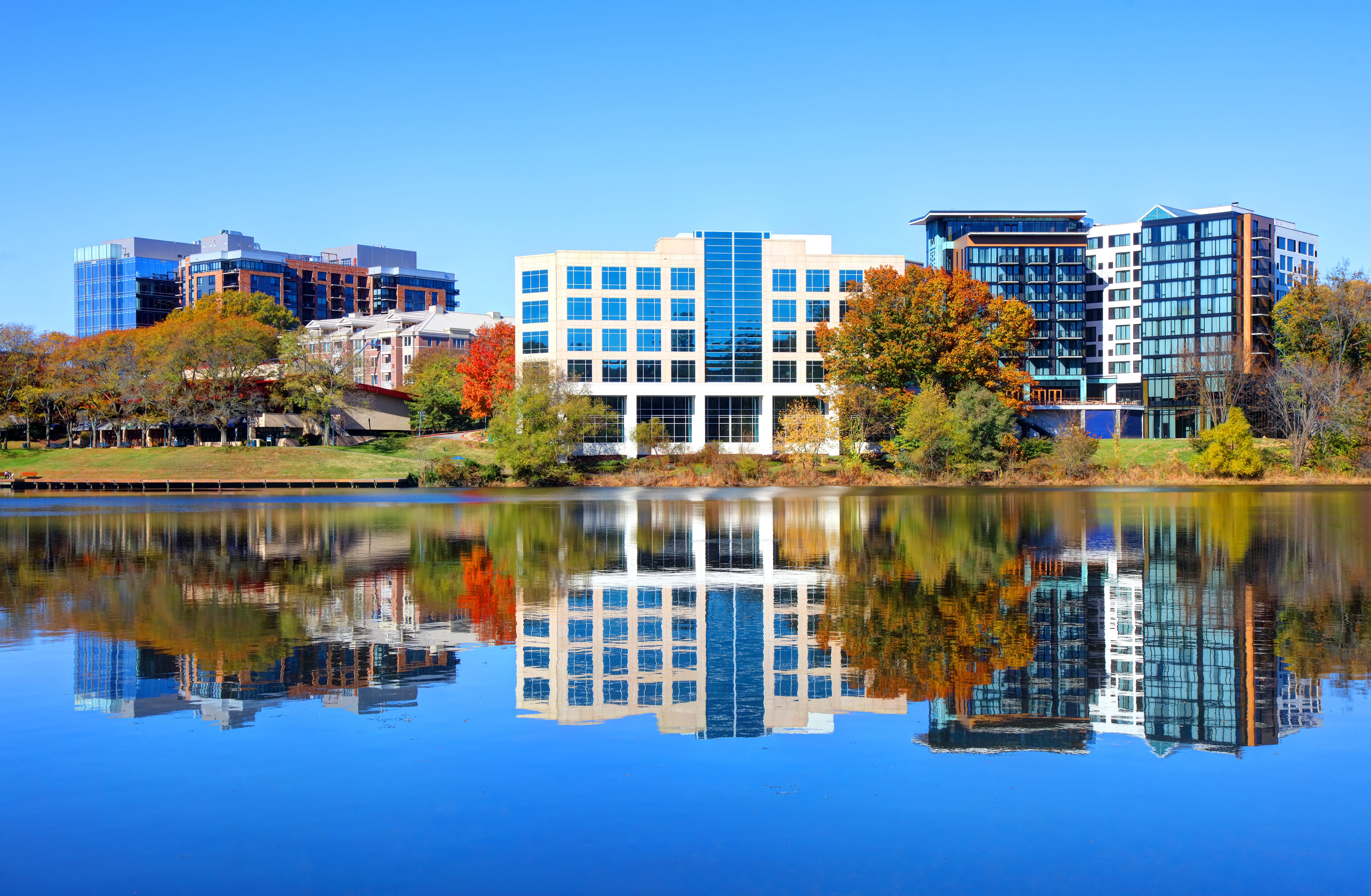 Modern office and apartment buildings reflect in a calm lake beside autumn trees under a clear blue sky.