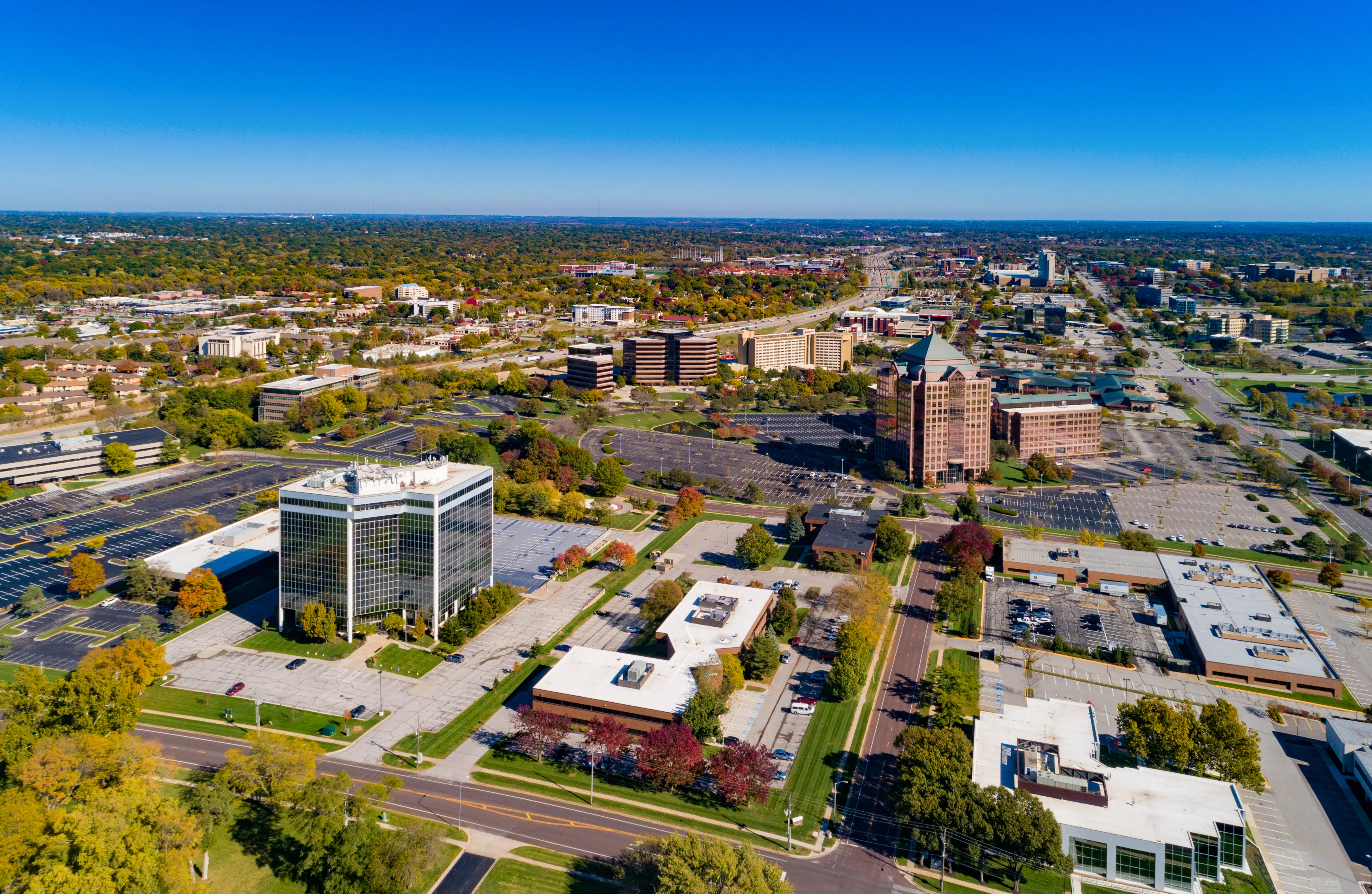 Aerial view of an office district with mid-rise buildings, parking lots, roads, trees, and a clear blue sky.