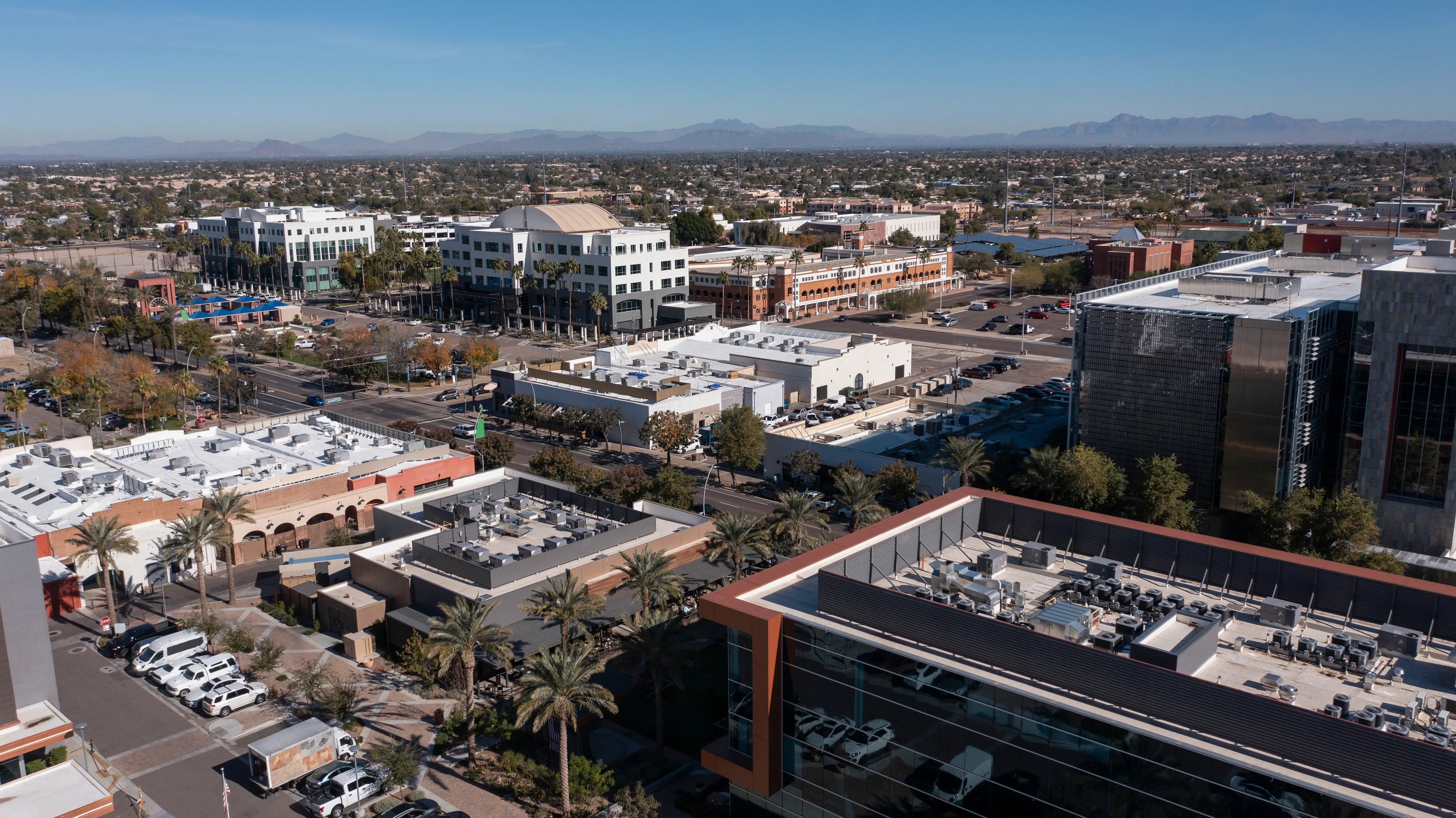 Aerial view of a sunny urban business district with office buildings, streets, parking lots, palm trees, and distant mountains.