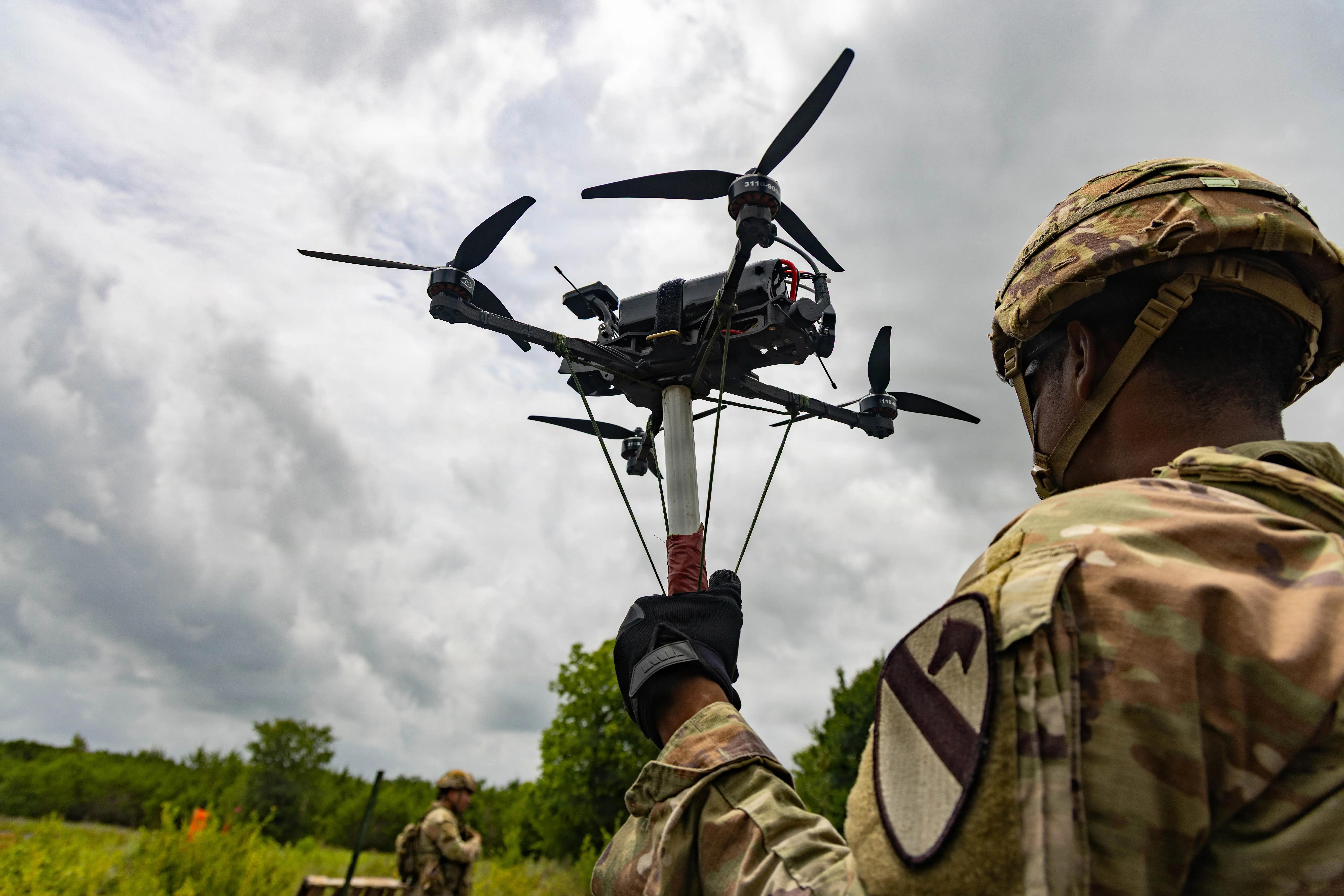 A soldier holds a small black quadcopter drone in a field with an overcast sky above.