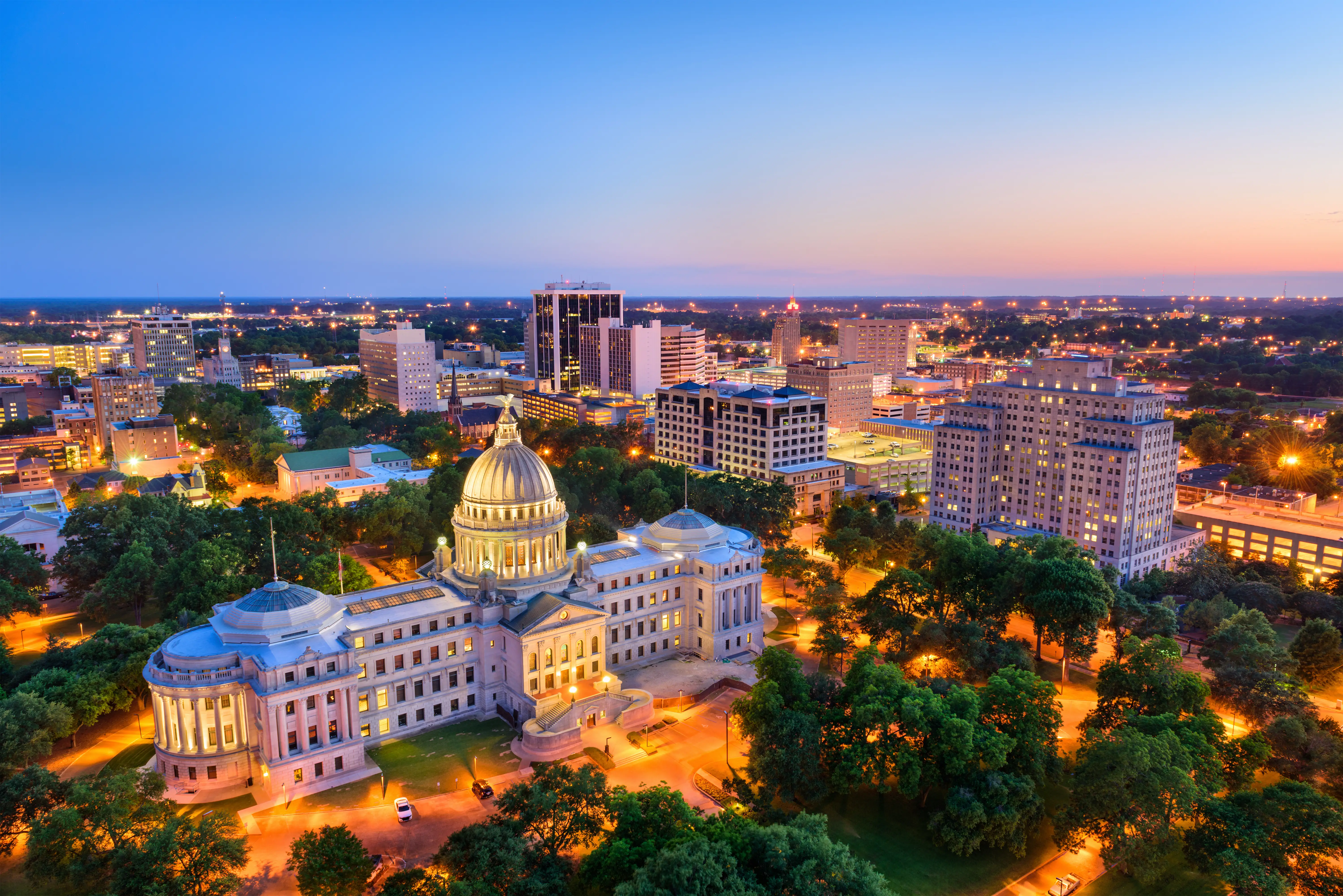 Illuminated Mississippi State Capitol stands amid trees and downtown Jackson skyline at dusk.