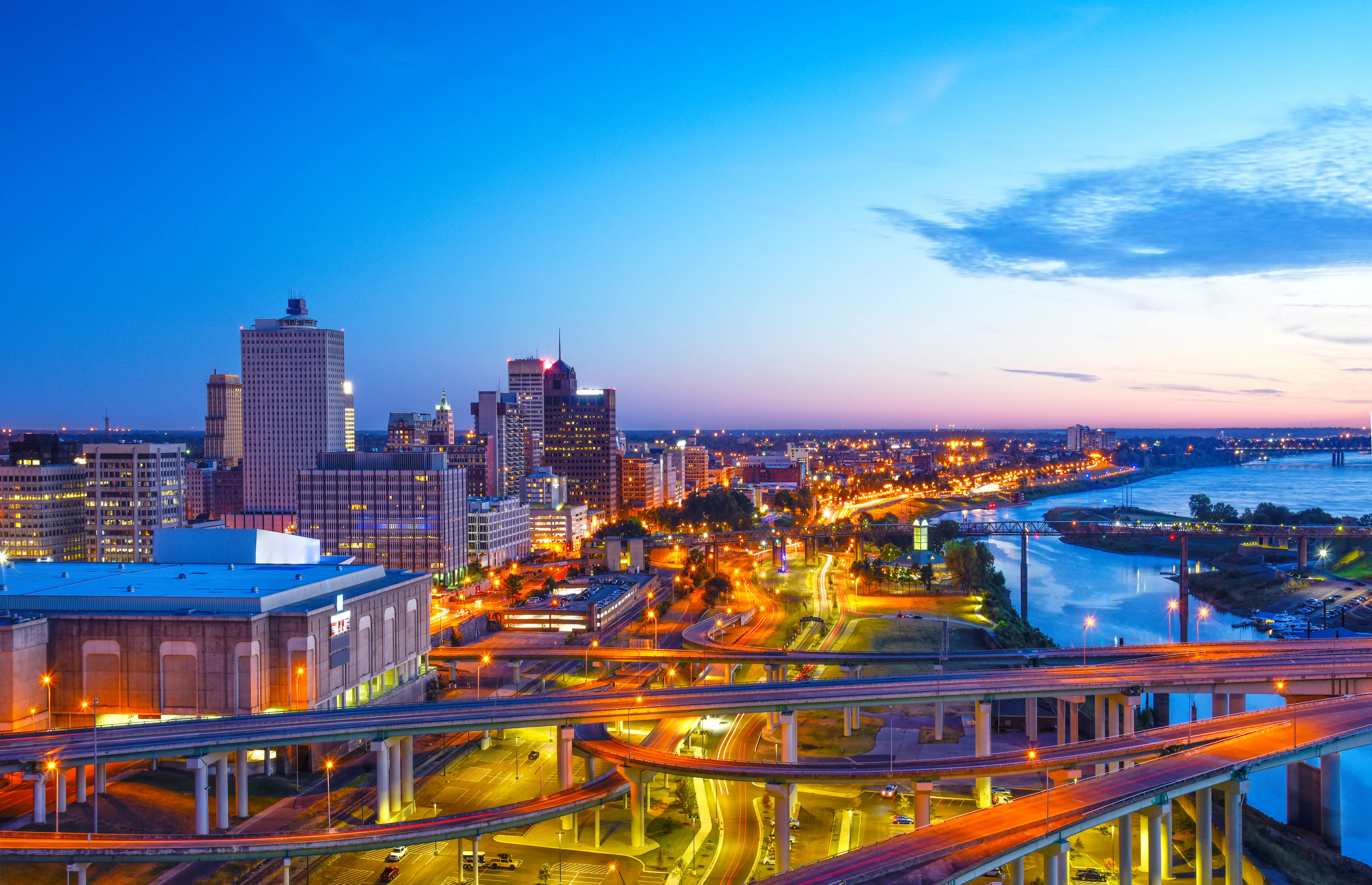 Downtown Memphis skyline and illuminated highway interchanges beside the Mississippi River at dusk.