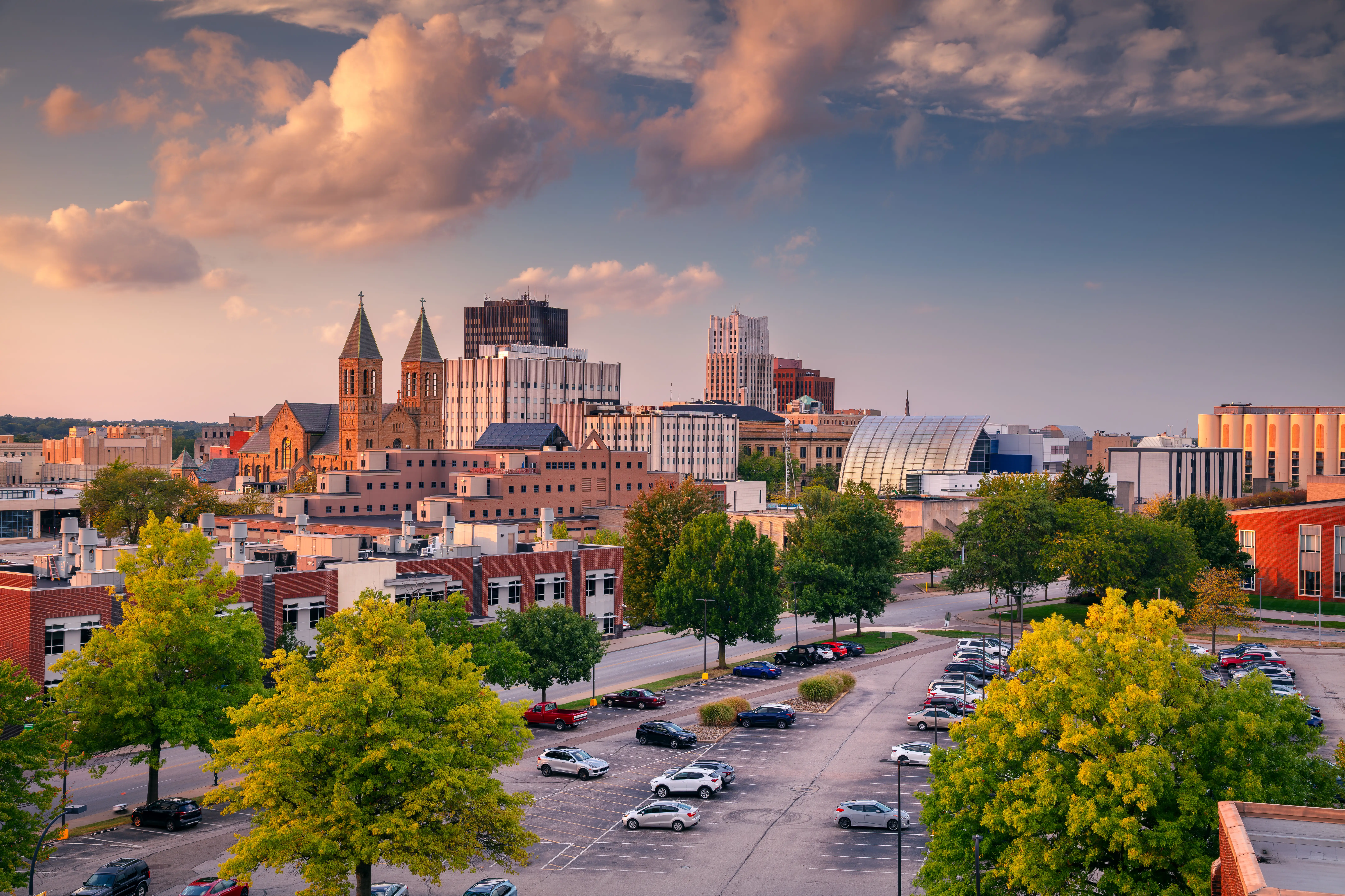 Downtown Fort Wayne skyline rises beyond a tree-lined parking lot under warm sunset clouds.