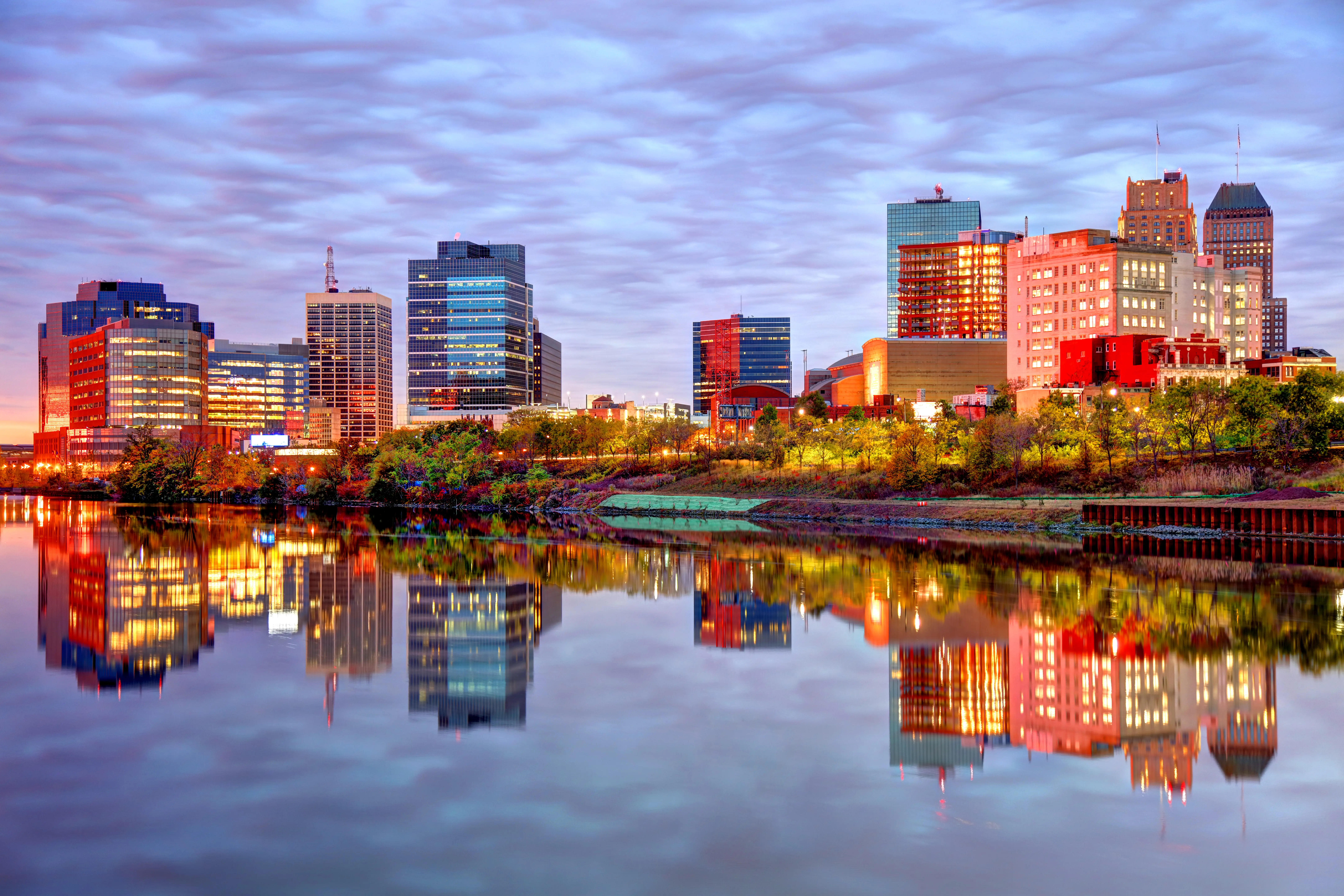 Illuminated downtown skyline reflects across a calm river at dusk under a cloud-filled sky.