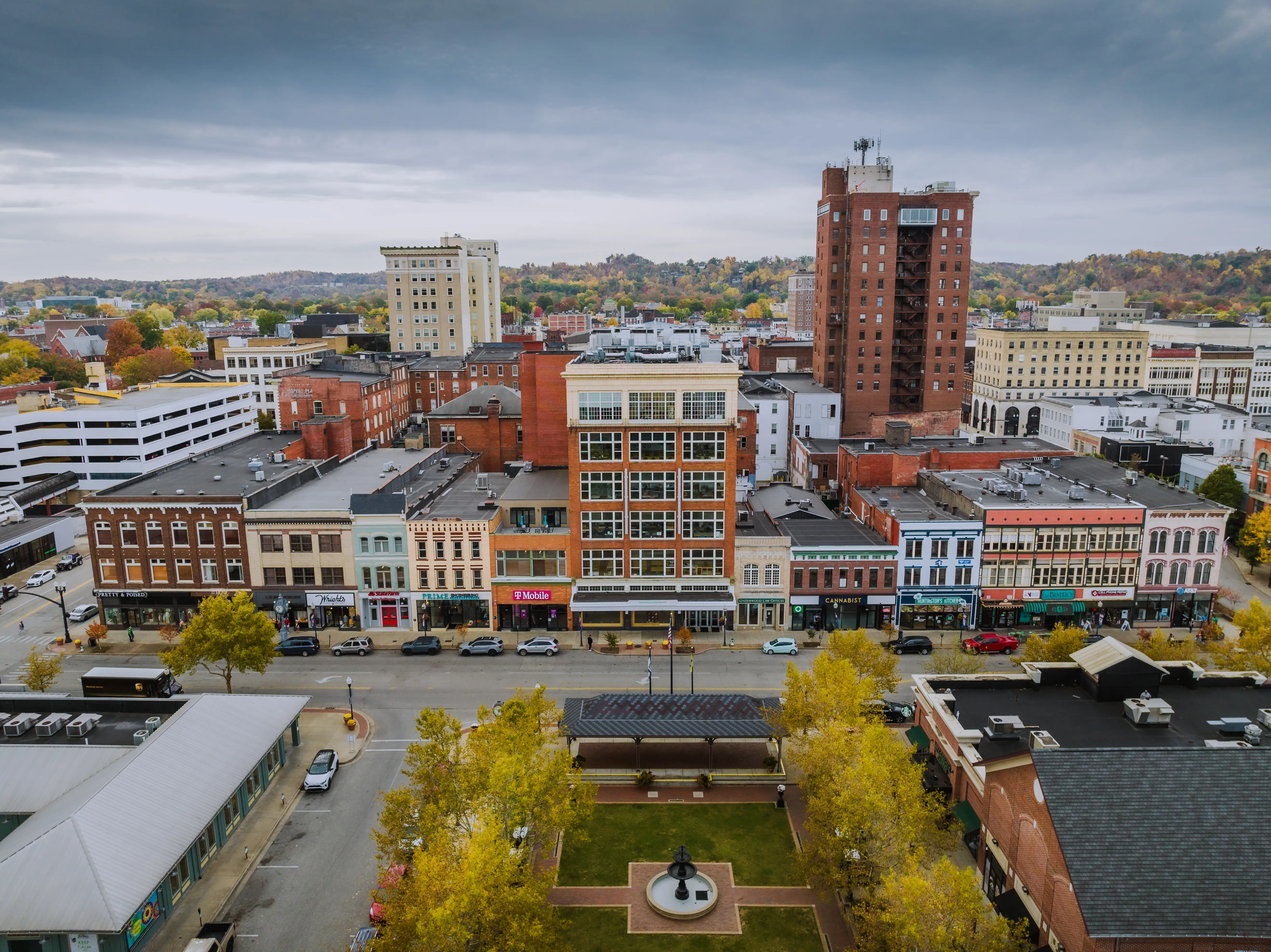 Downtown streetscape with storefronts, mid-rise buildings, a small park, and autumn trees under a cloudy sky.