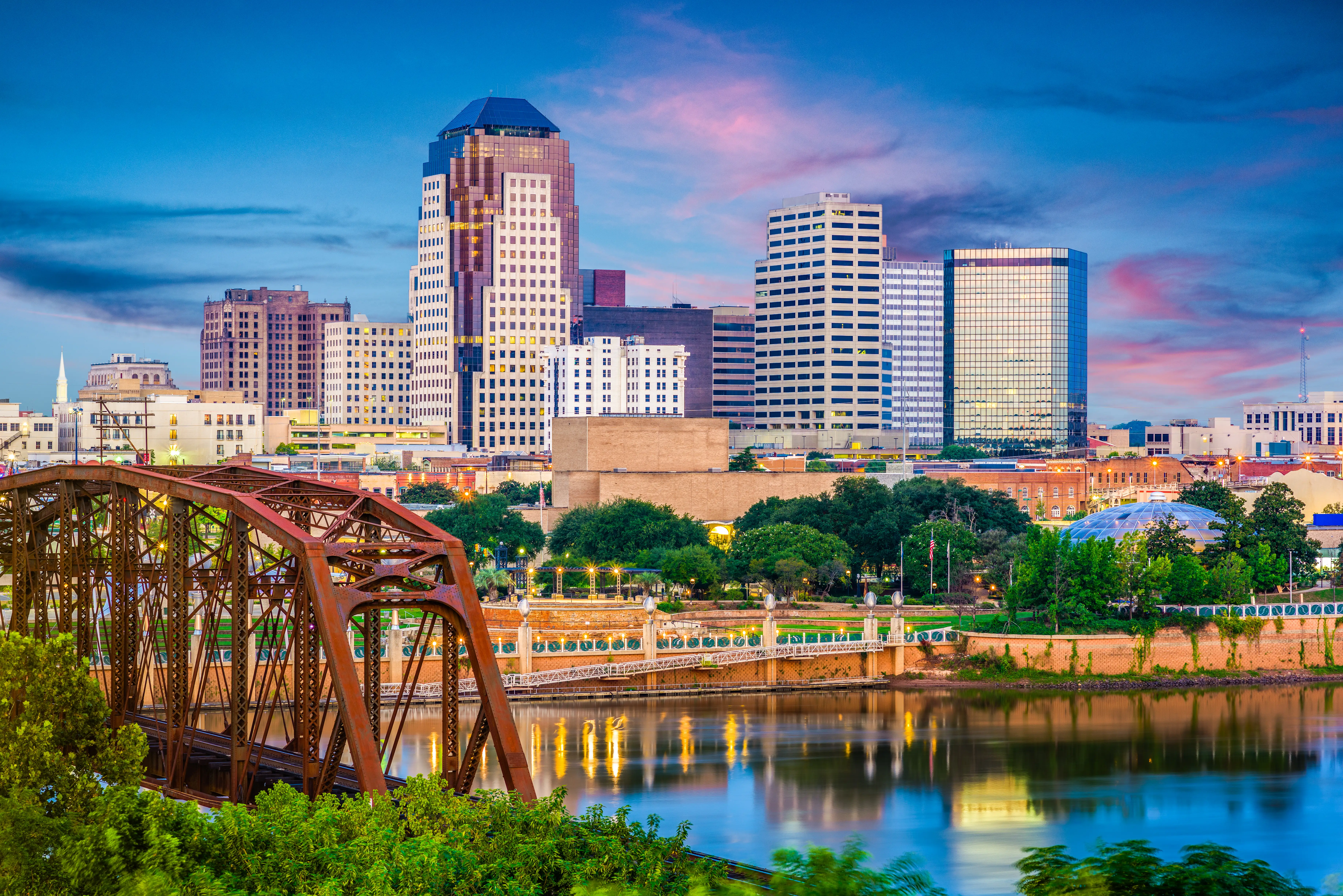 Downtown Shreveport skyline rises behind a truss bridge and river at dusk with colorful reflected lights.