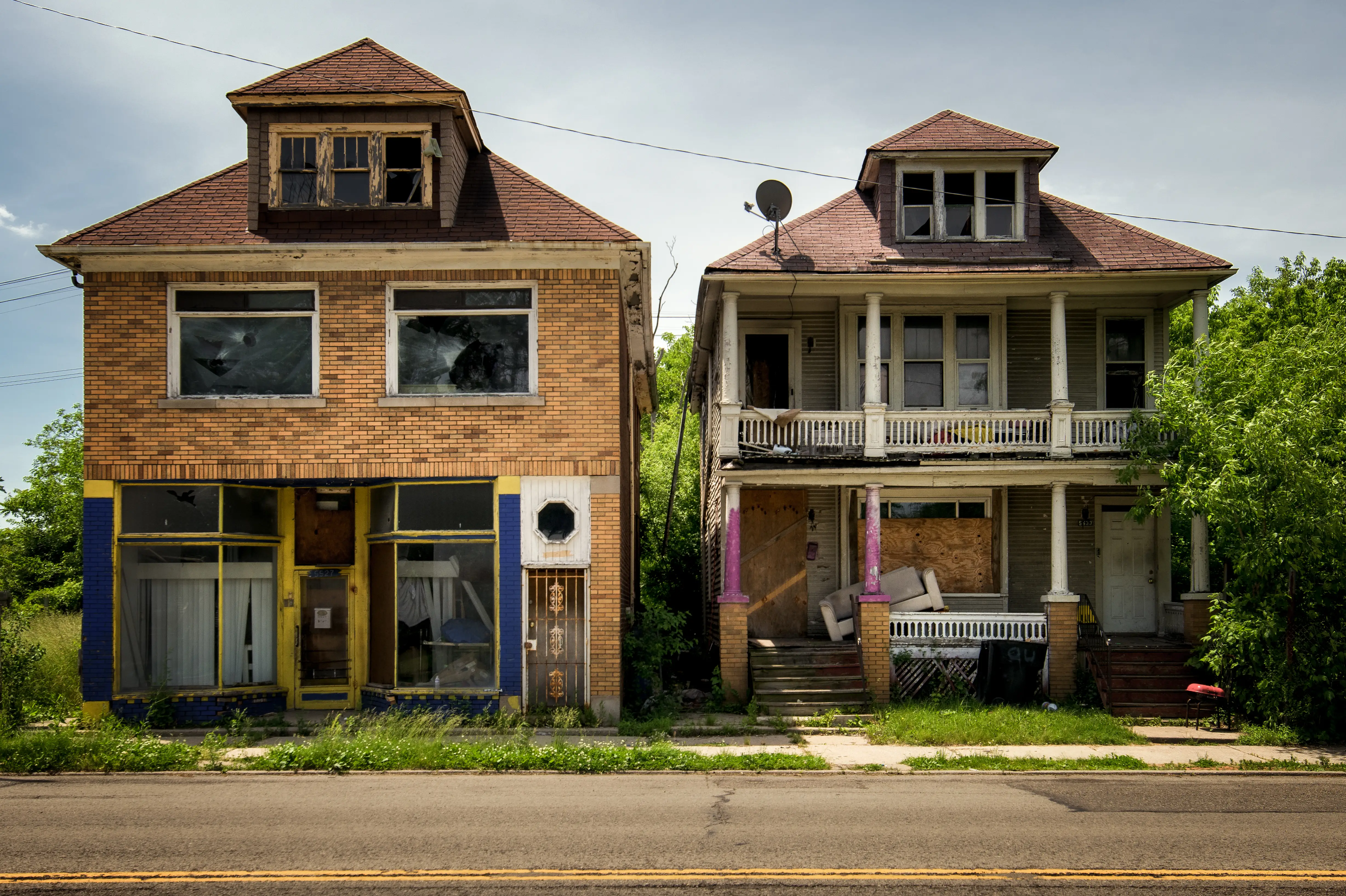 Two weathered vacant houses with broken windows and overgrown yards stand along a quiet urban street.