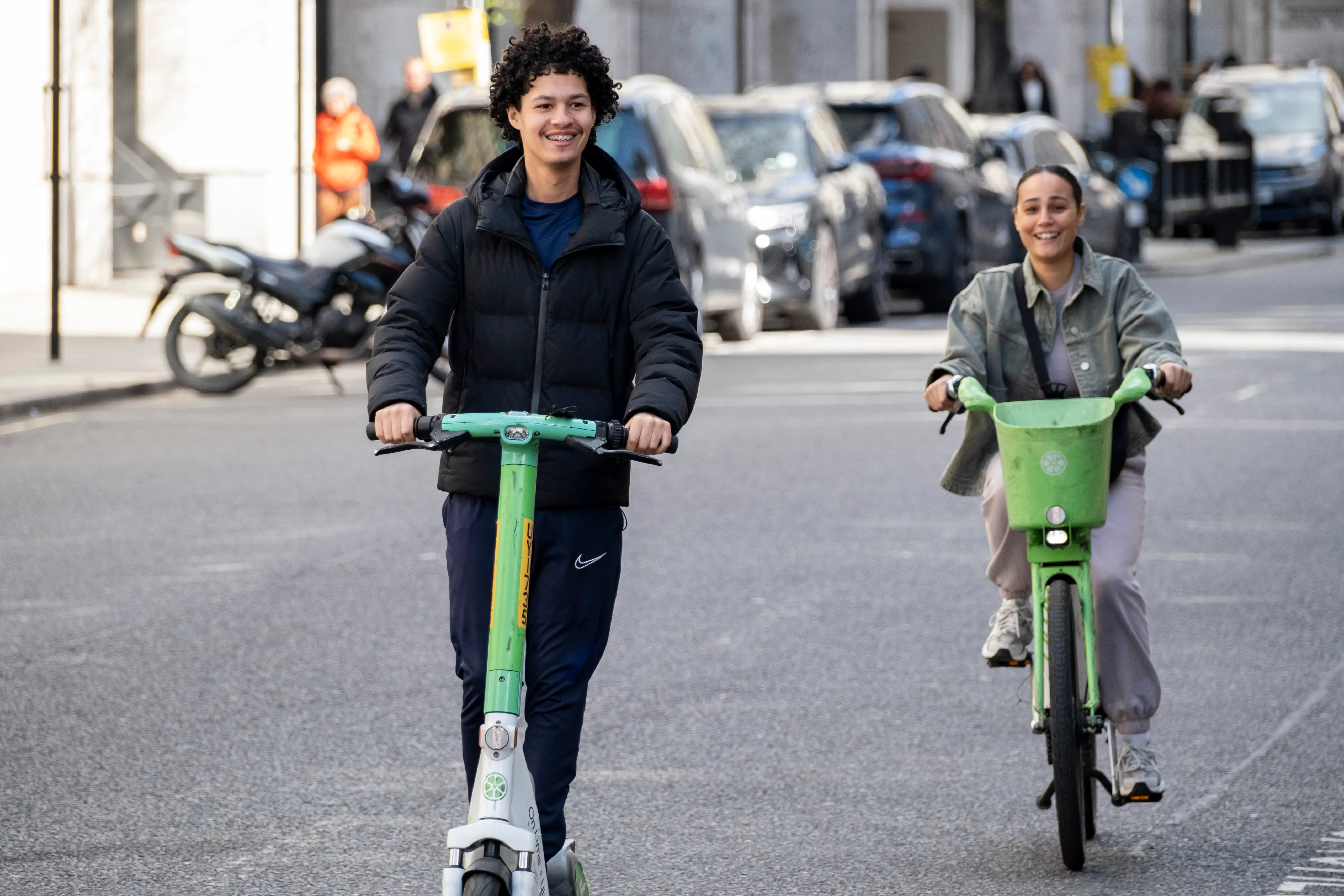 A man rides a Lime scooter as a woman next to him rides a Lime bicycle.