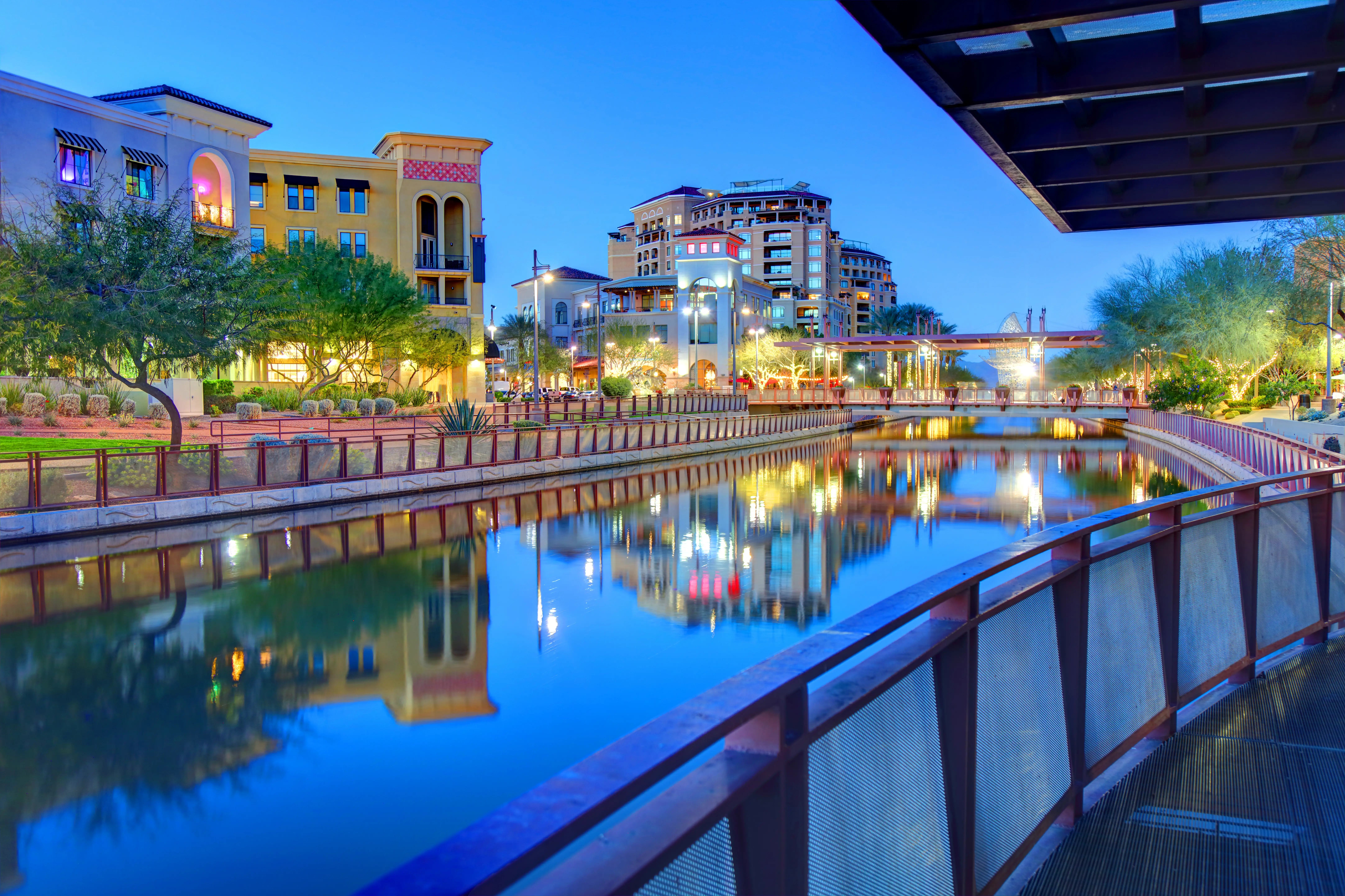 Illuminated buildings and pedestrian railings reflect in a calm urban canal at blue hour.