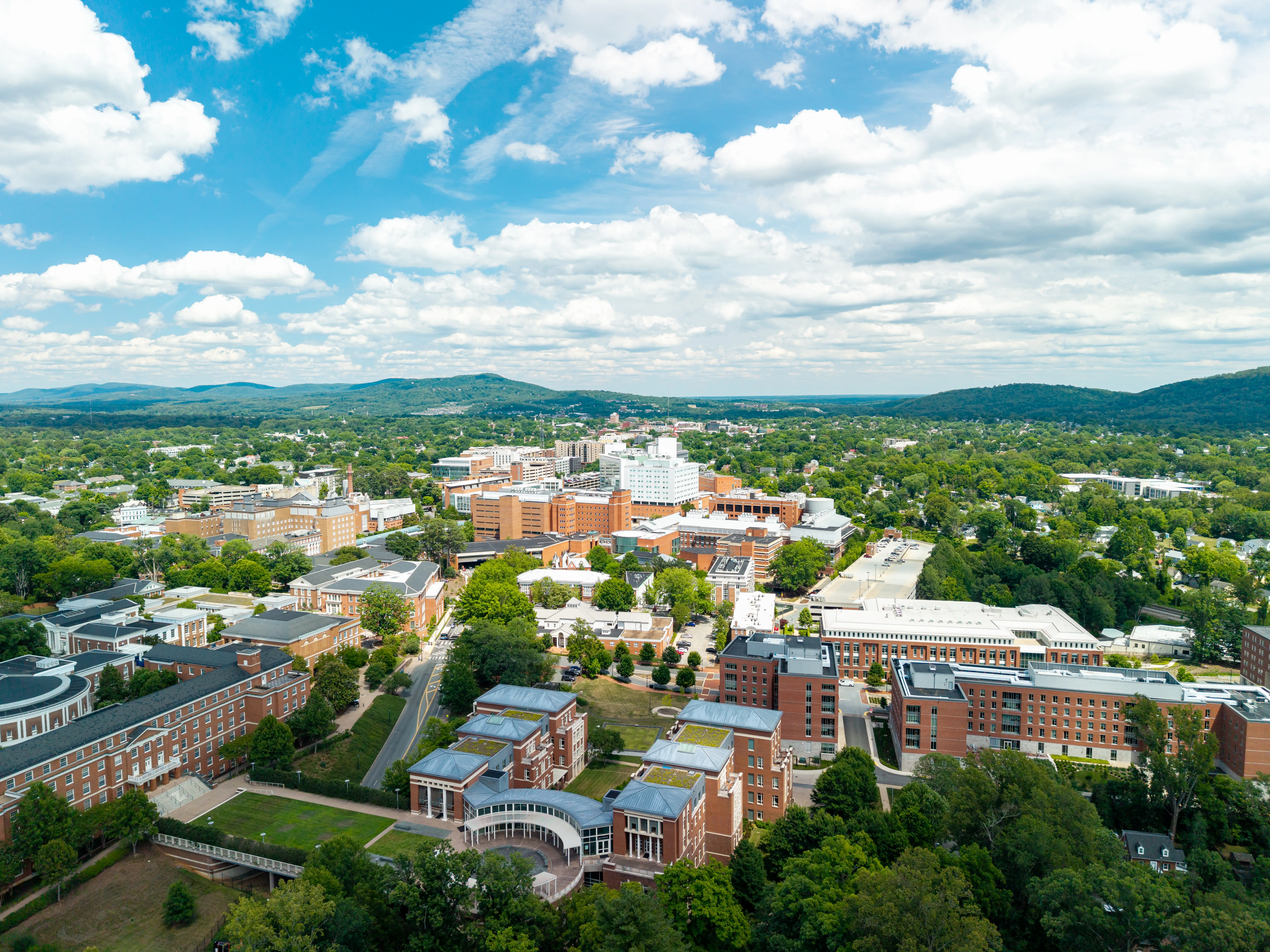 Charlottesville, Virginia, wide aerial drone view looking over the expansive University of Virginia campus and medical center complex, stretching toward rolling green hills under a bright sky with scattered clouds.
