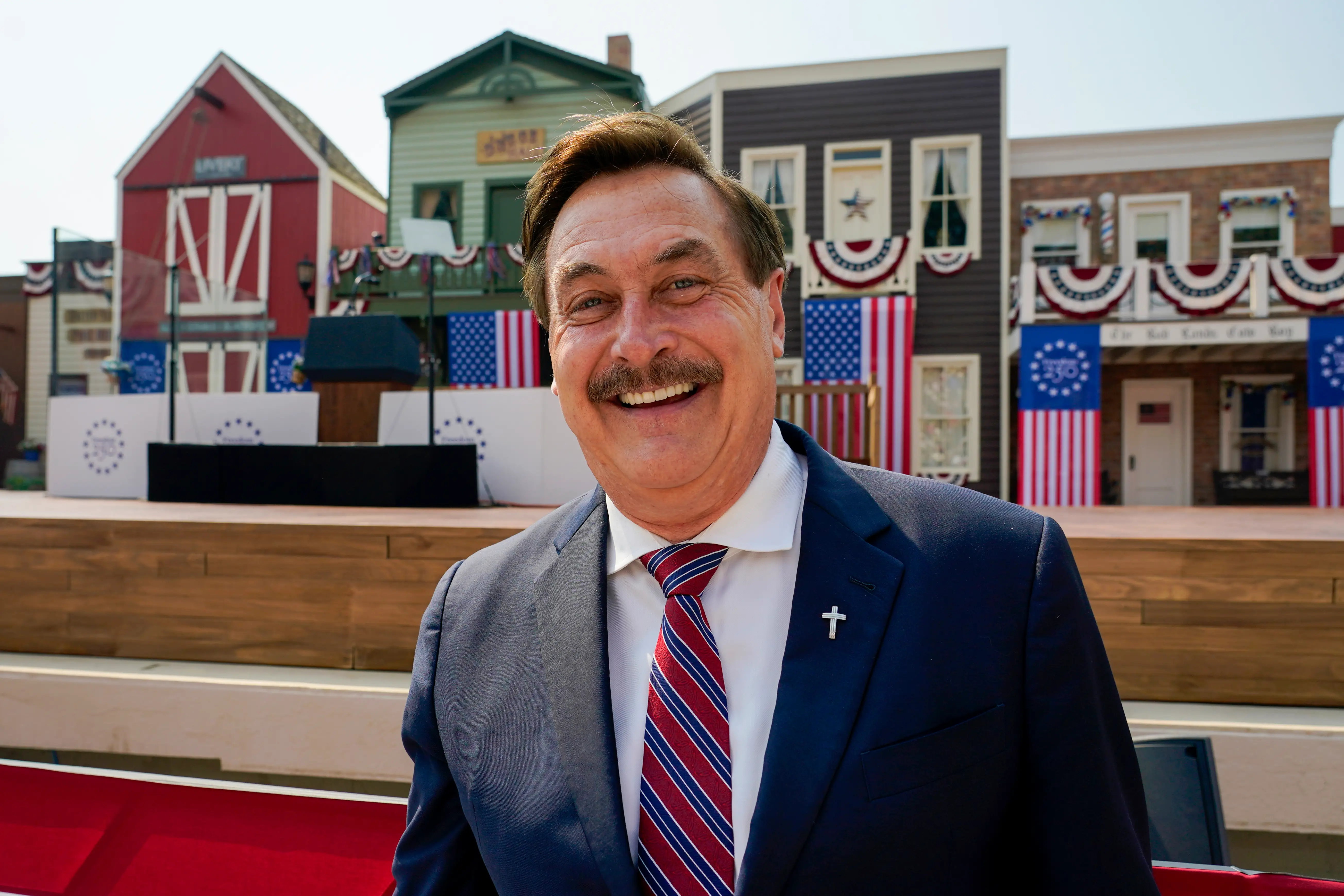 Mike Lindell smiles in a suit before a patriotic stage set with American flags and bunting.