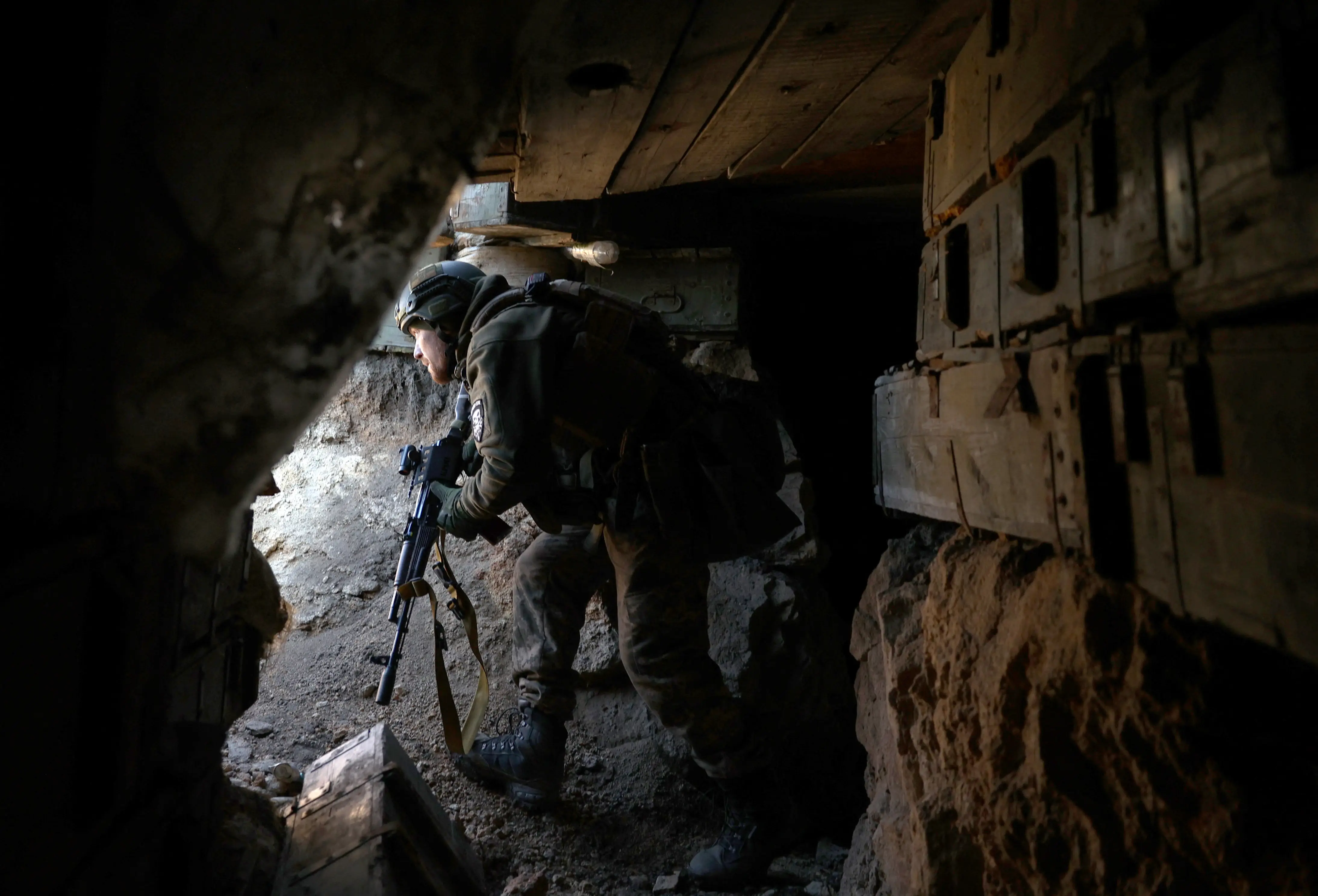 A camouflaged armed person crouches inside a dark fortified position beside rough concrete and wooden supports.