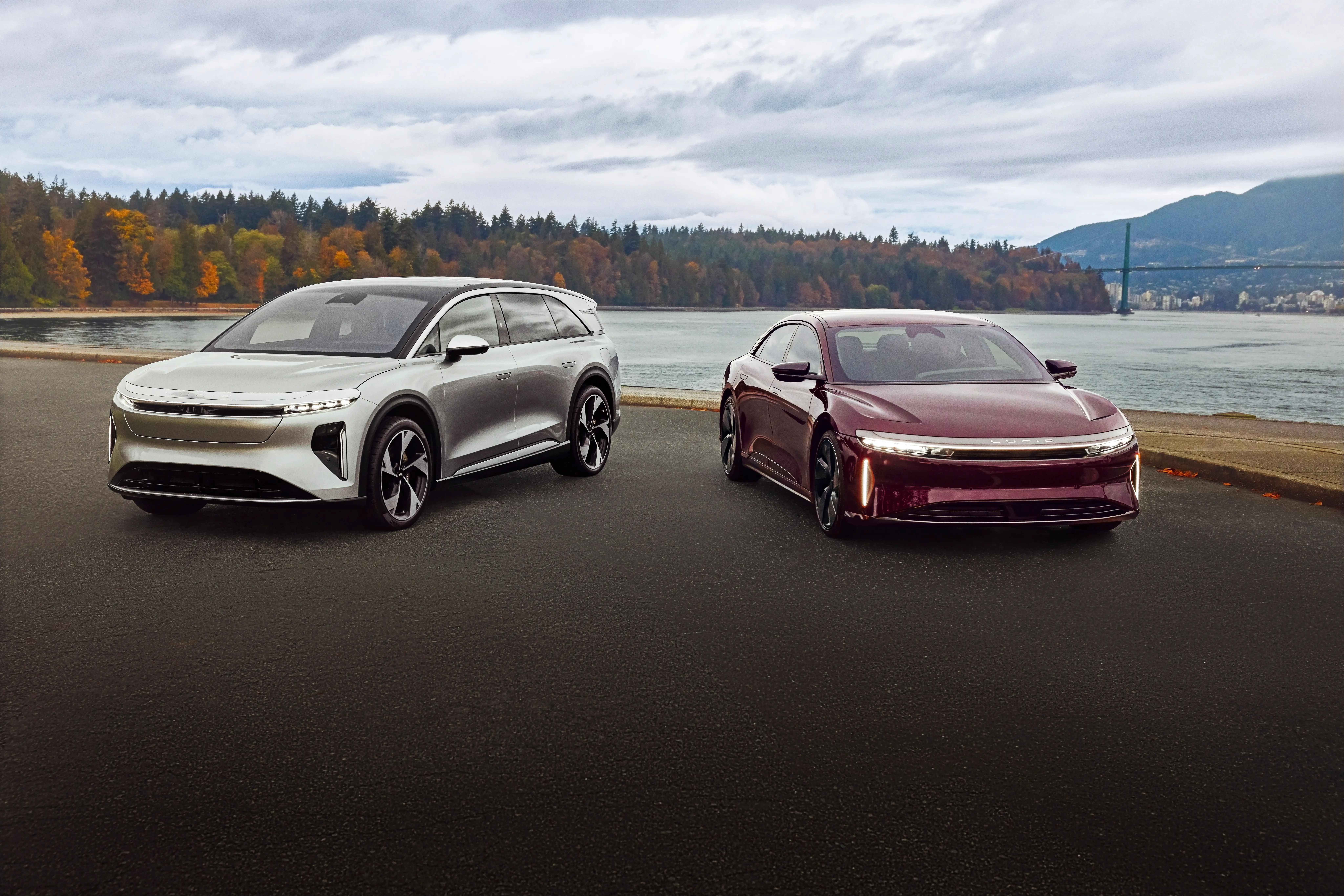 A silver Lucid Gravity SUV is parked next to a red Lucid Air sedan. They're both in a parking lot that's close to a body of water.