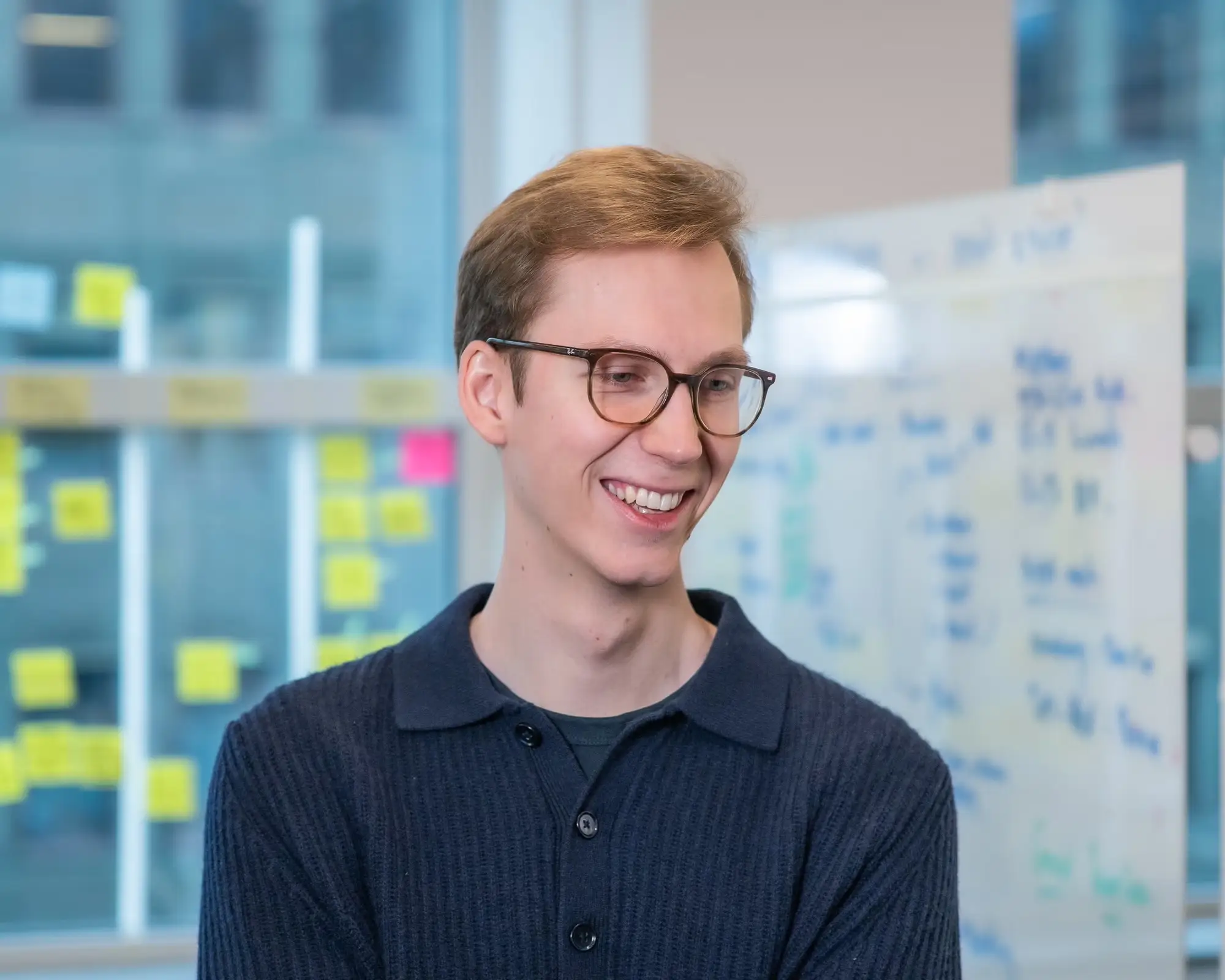 Smiling person wearing glasses stands in an office beside a whiteboard and colorful sticky notes.