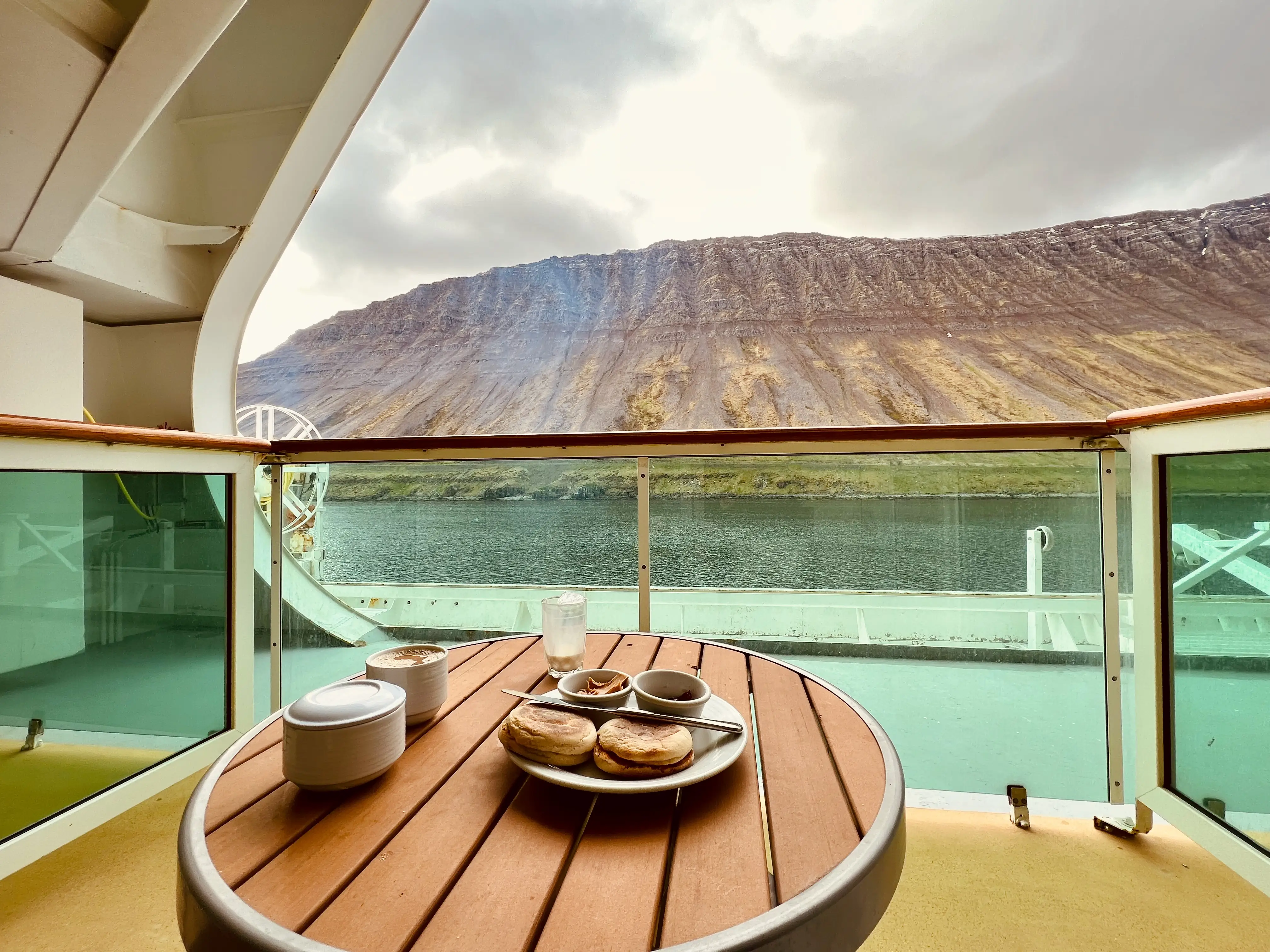 Plate with breakfast and coffee mug on table of cruise room balcony in front of views of rocky structure, water