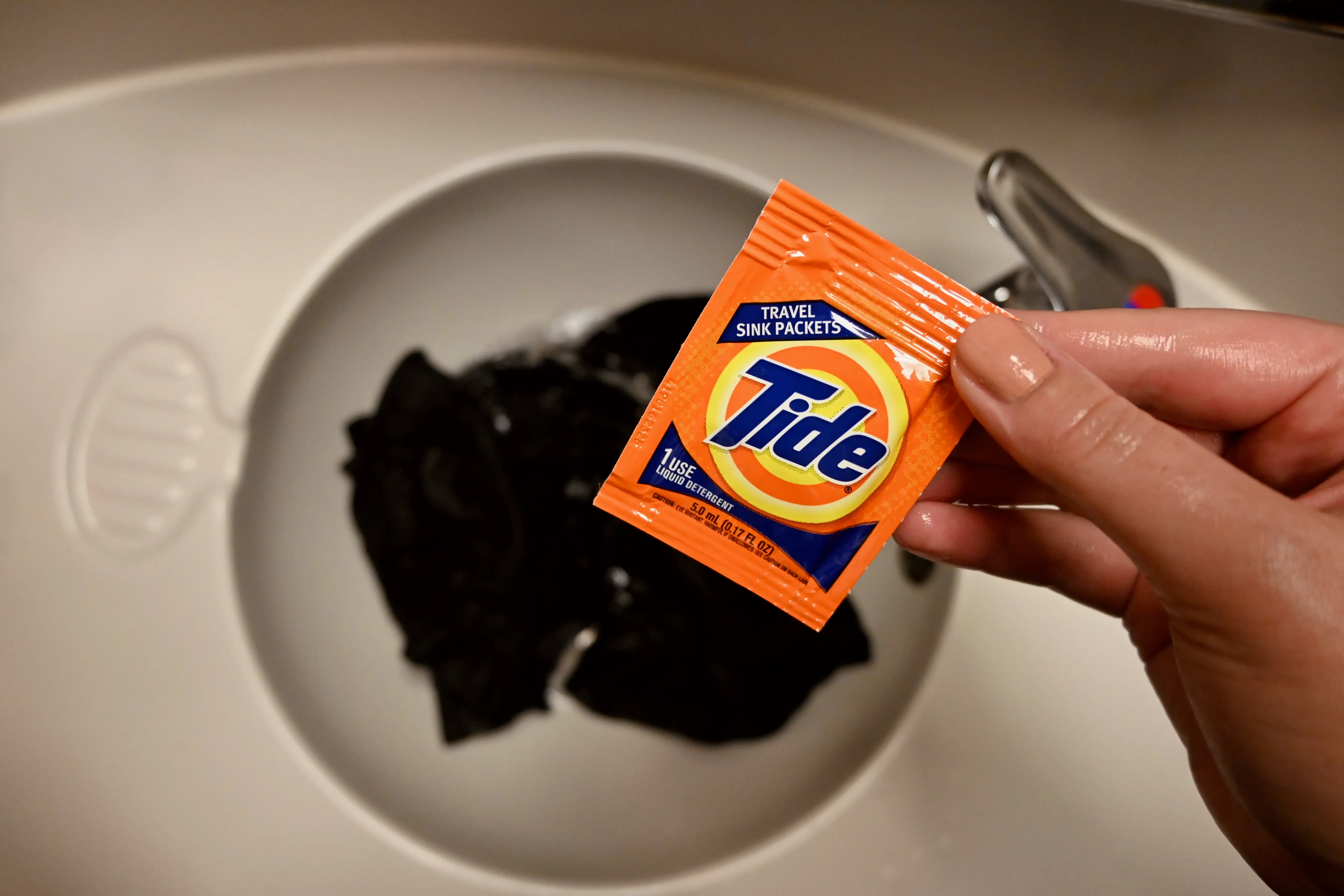 Hand holding a Tide travel detergent packet over a sink with clothes in it