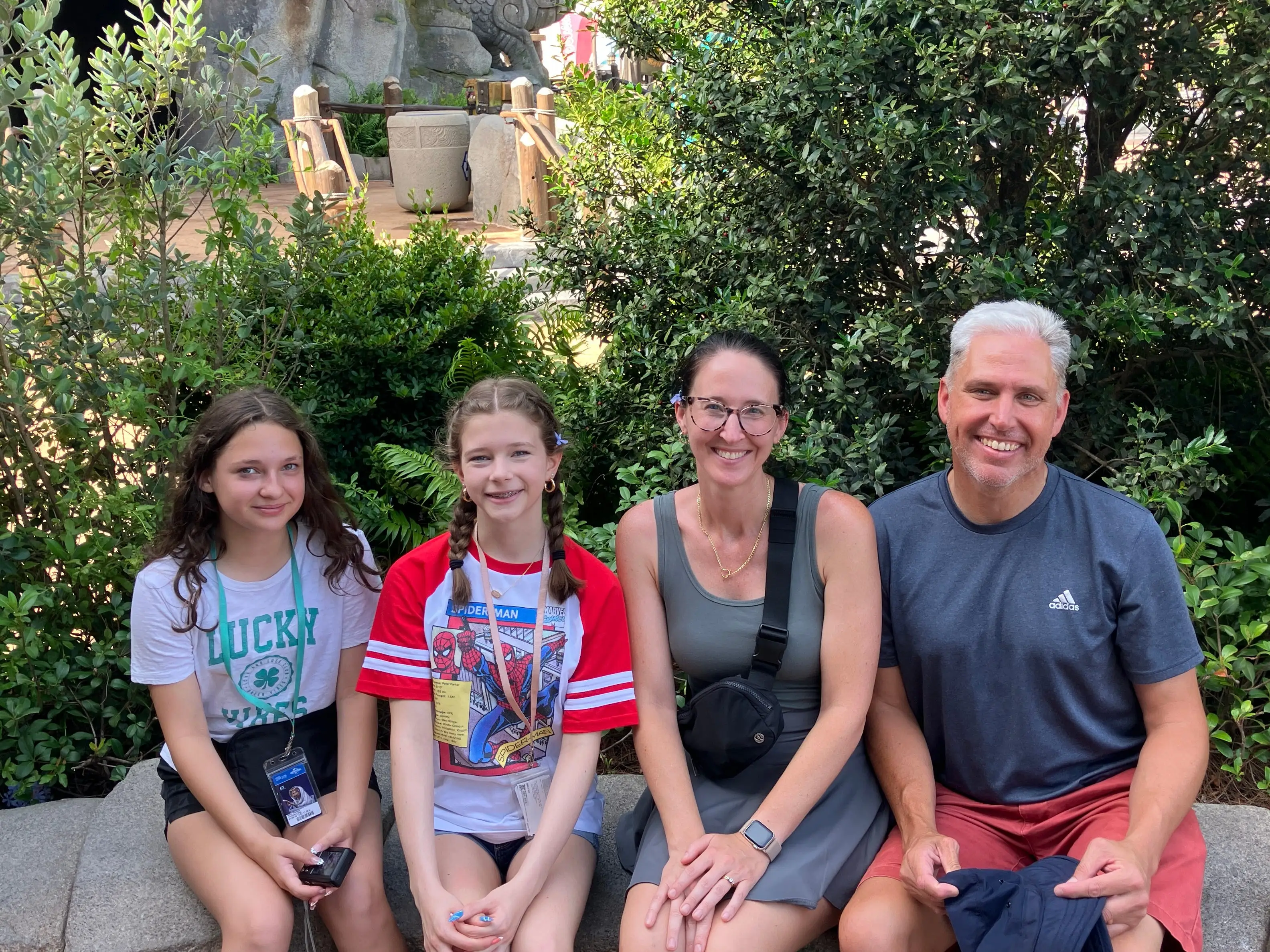 Four visitors sit on a stone wall among greenery near a rocky themed cave entrance.
