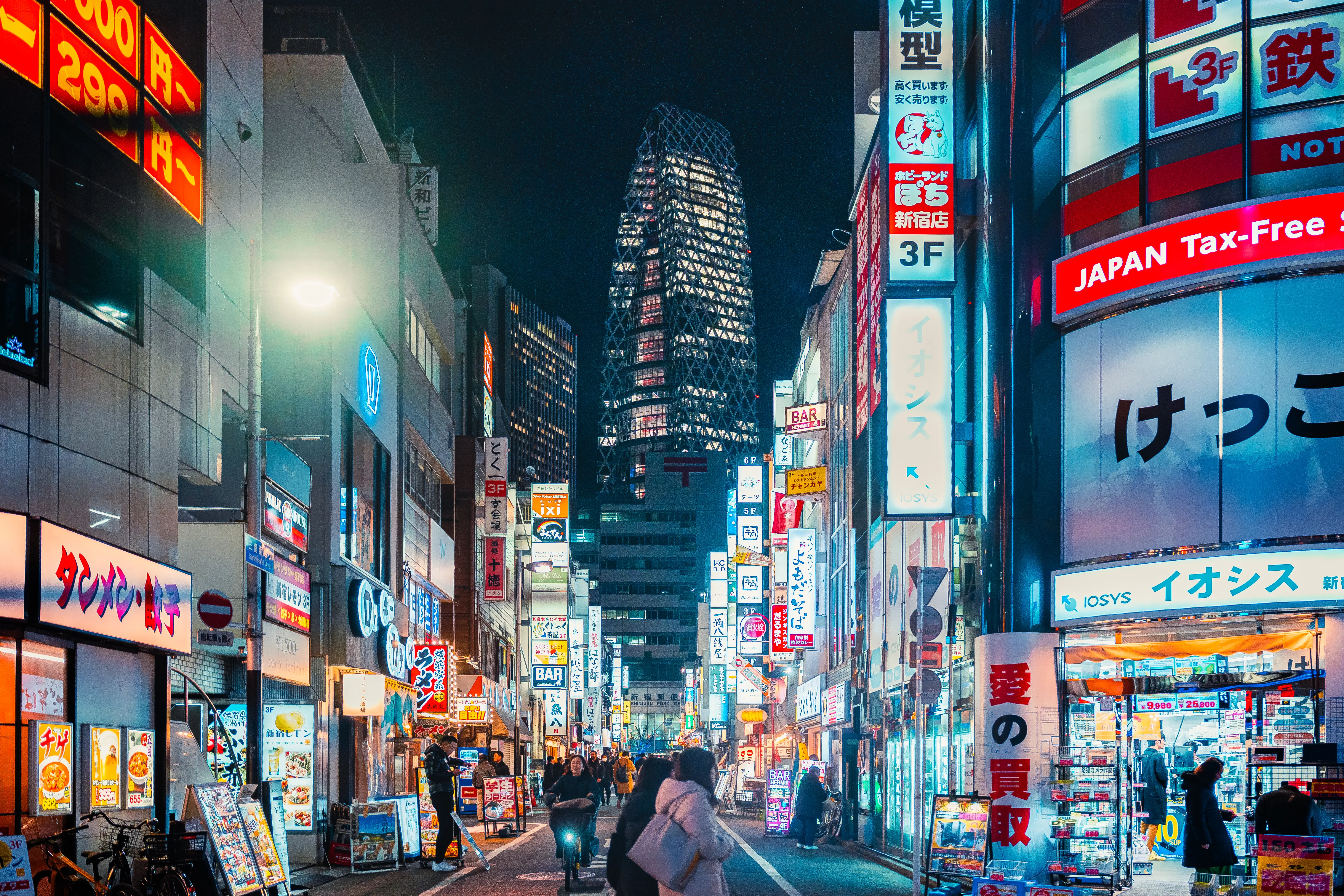 Neon-lit Japanese shopping street at night with storefront signs, pedestrians, and a tall illuminated tower in the background.